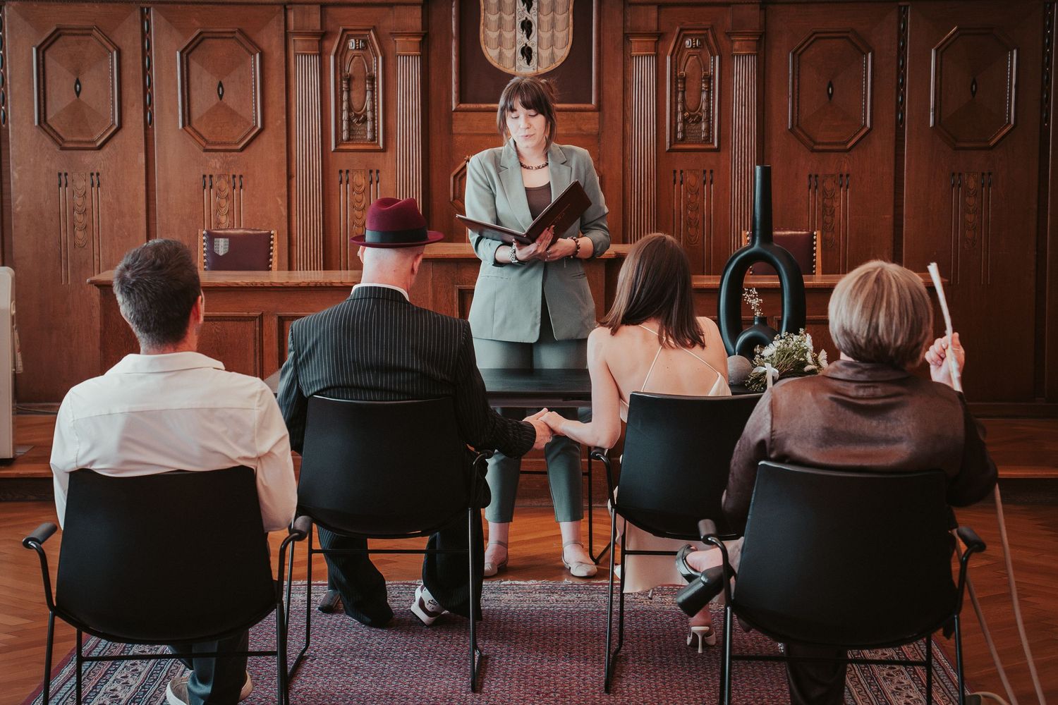 A celebrant reads during an intimate small wedding ceremony inside a wood-paneled room with seated guests.