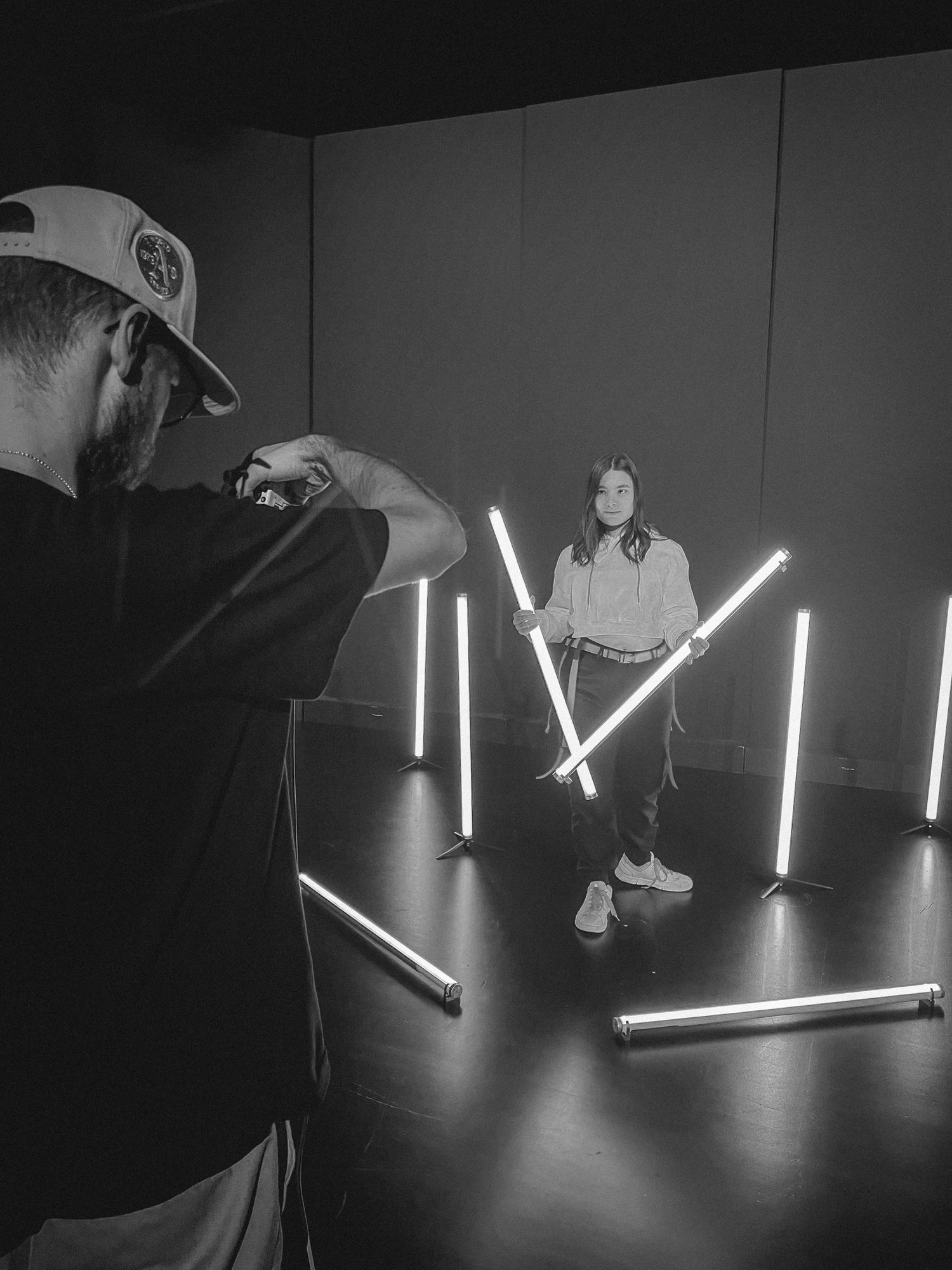 Photographer capturing a woman posing with neon light tubes in a dark studio, black and white.