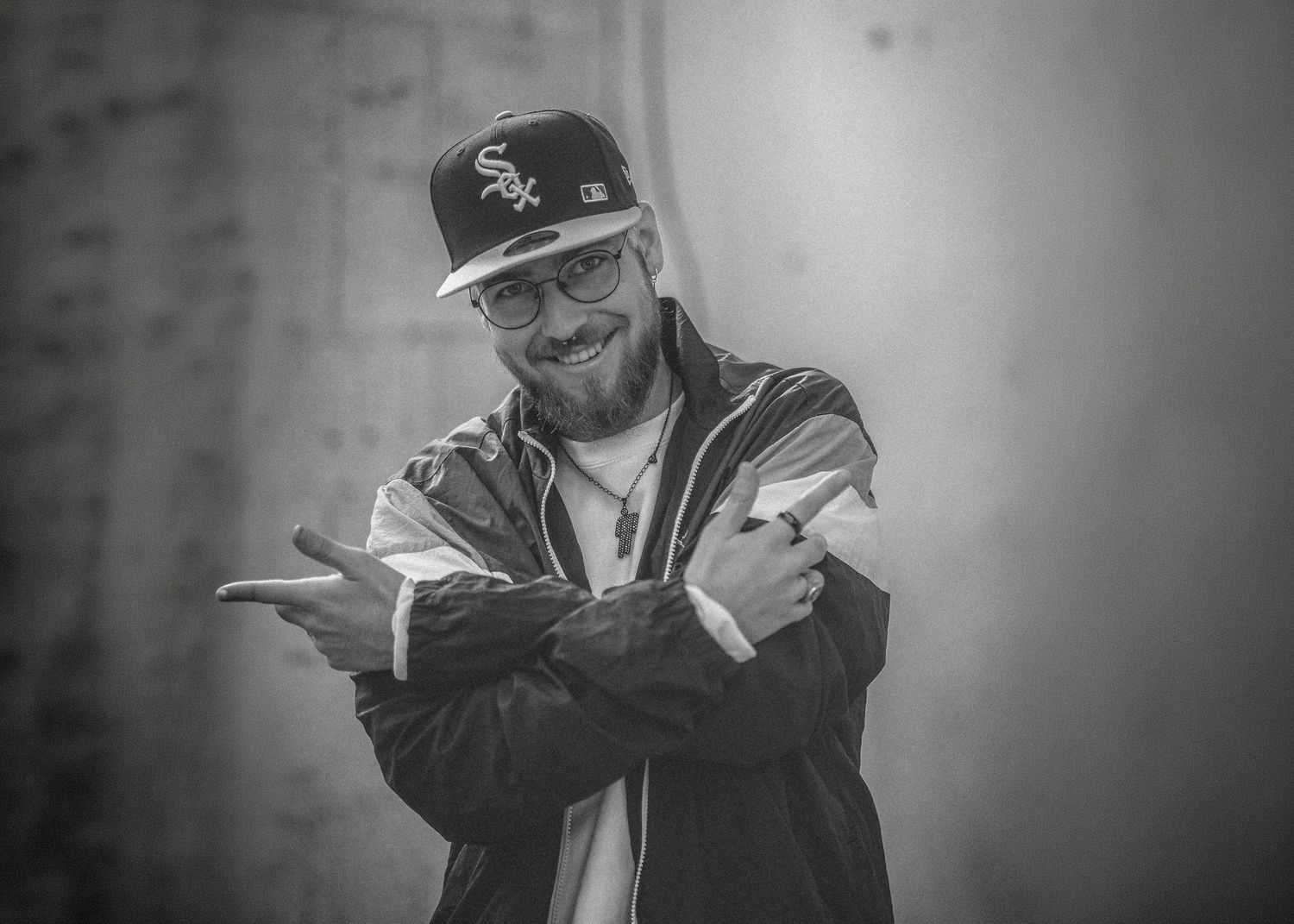 Smiling bearded man in baseball cap pointing fingers forward in black and white portrait.