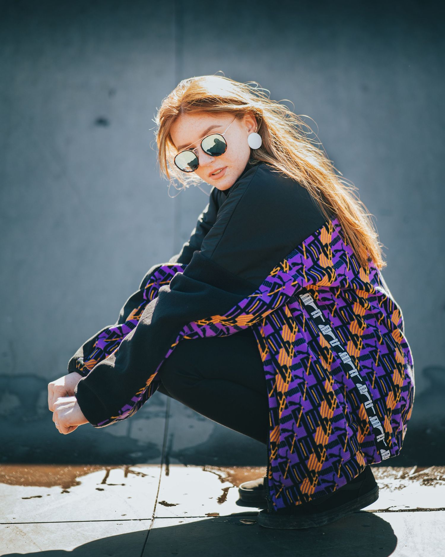Redhead woman in purple patterned jacket with mirrored sunglasses crouching against gray wall.