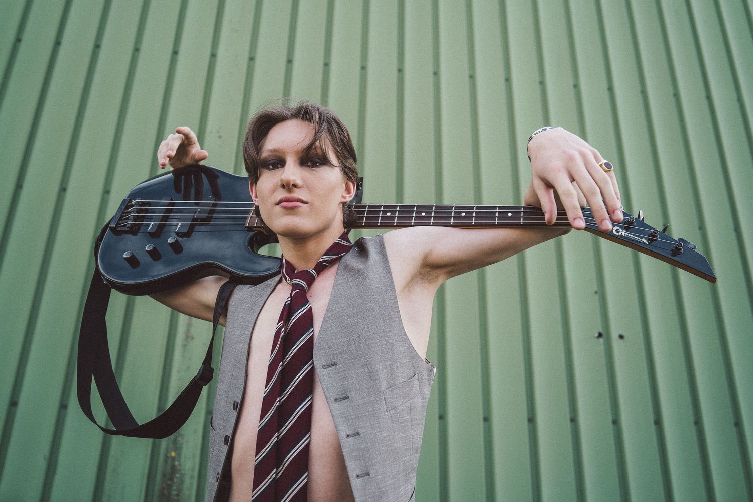 Young musician posing with black bass guitar on shoulders against green corrugated metal wall wearing tie and vest.