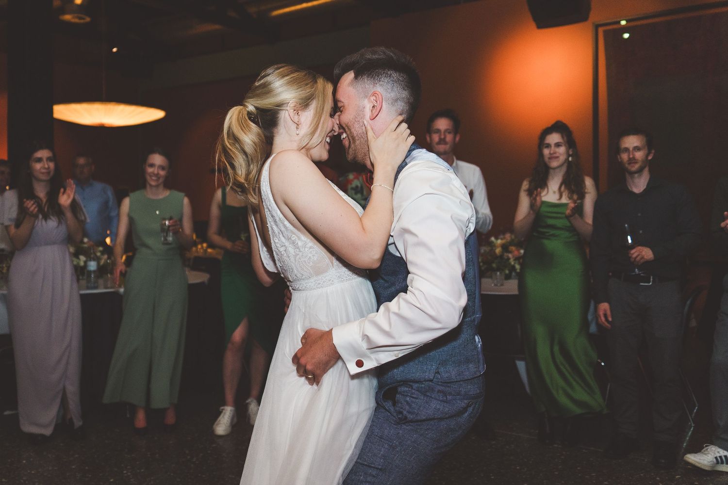 Bride and groom sharing first dance at wedding reception surrounded by guests in green dresses.