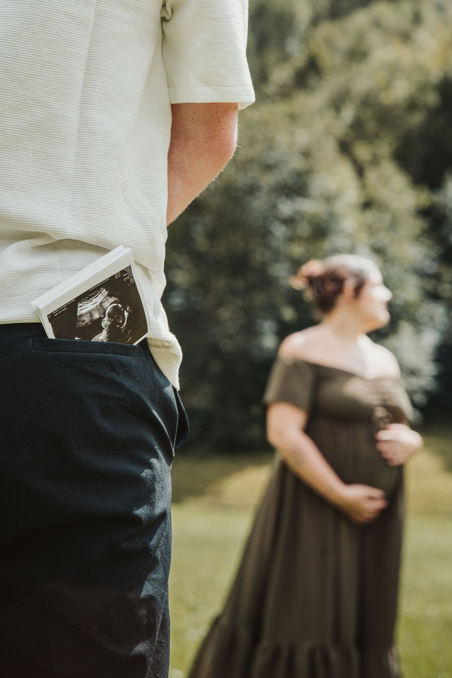 Man with ultrasound photo in pocket standing near pregnant woman in dark off-shoulder dress in a sunny park.