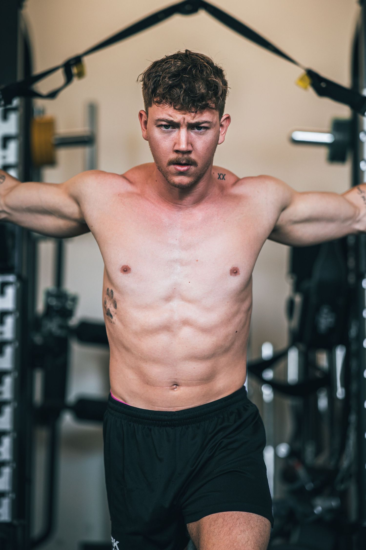 Muscular shirtless man performing cable fly exercise in a gym displaying defined physique and tattoos.