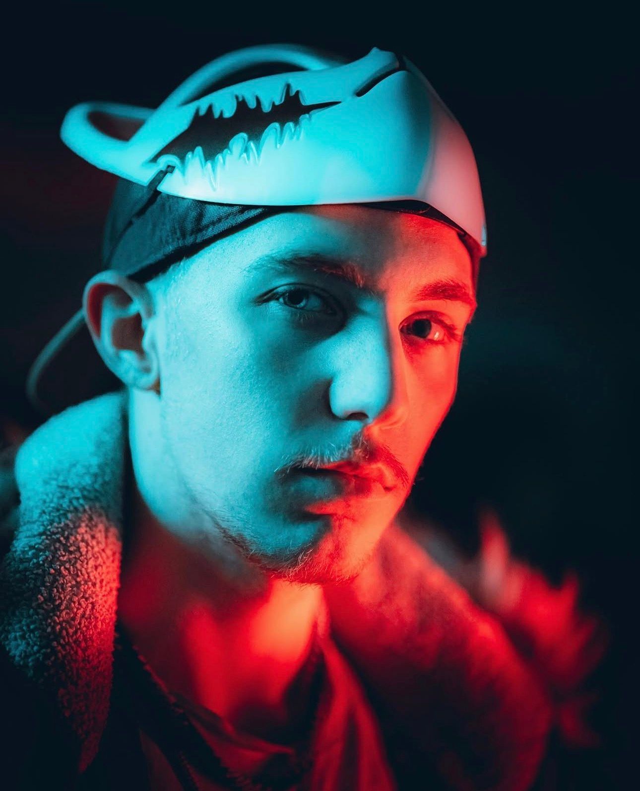 Young man wearing a cap illuminated by dramatic red and blue neon lights in a dark setting.
