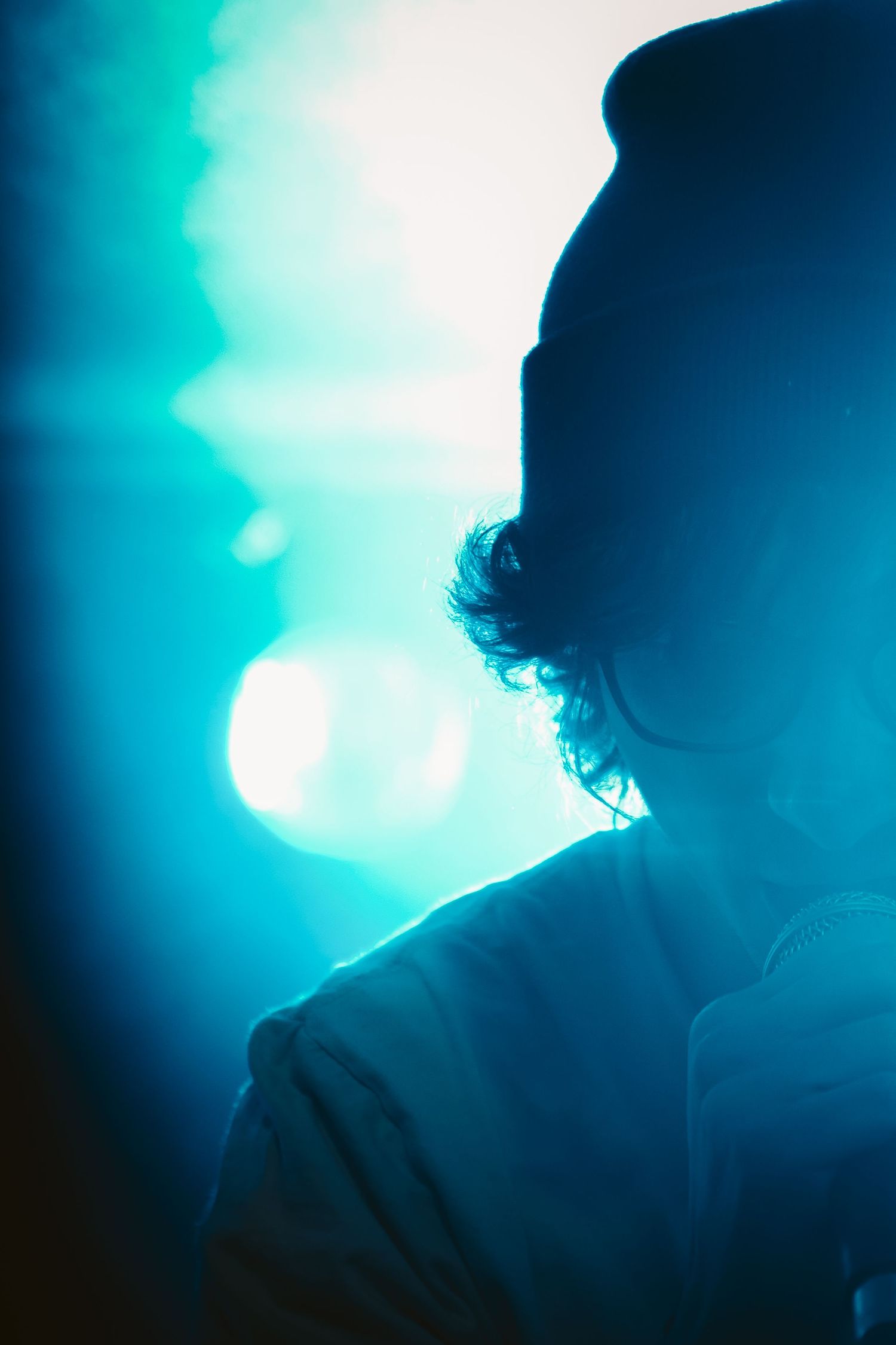 Silhouette of curly-haired musician against bright teal bokeh stage lights in moody concert shot.