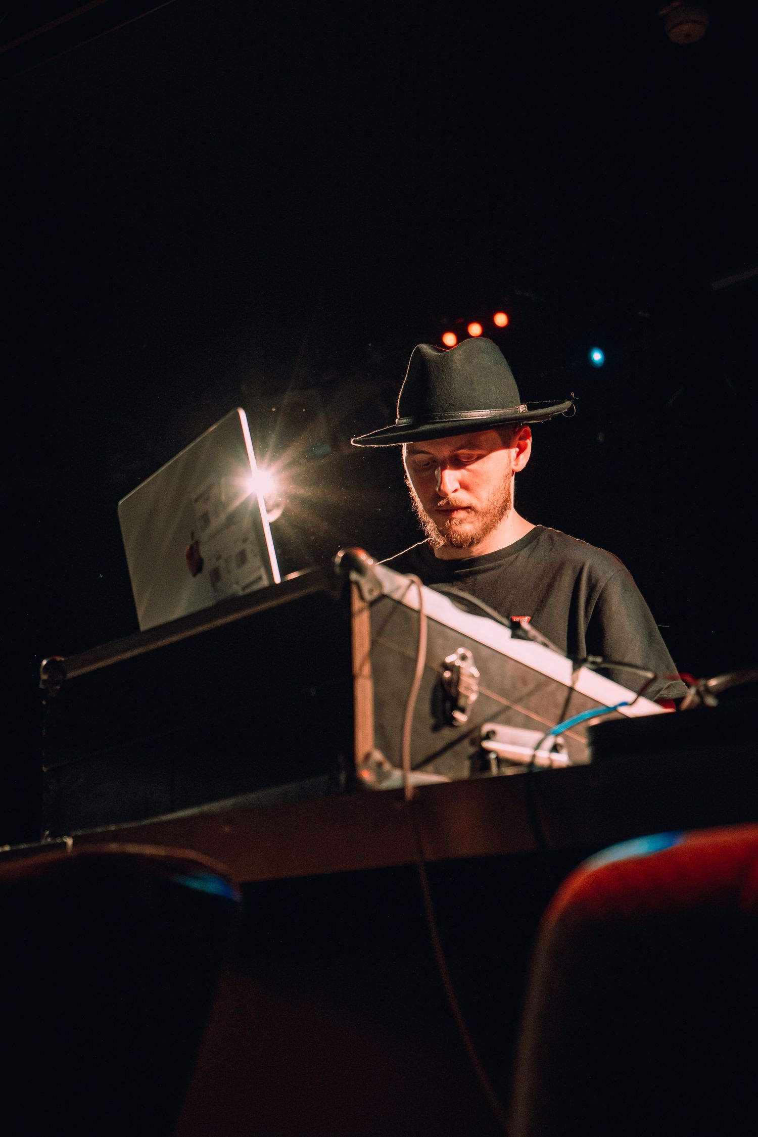 A bearded man wearing a wide-brim hat works at a laptop and equipment setup on a dimly lit stage.