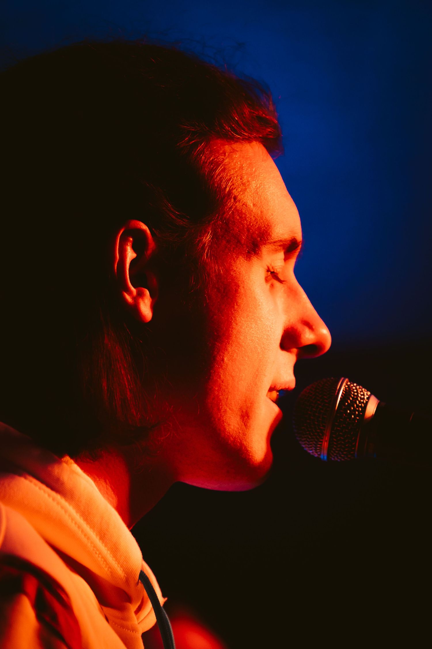 Close-up profile of male vocalist singing into microphone under warm red concert stage lighting.