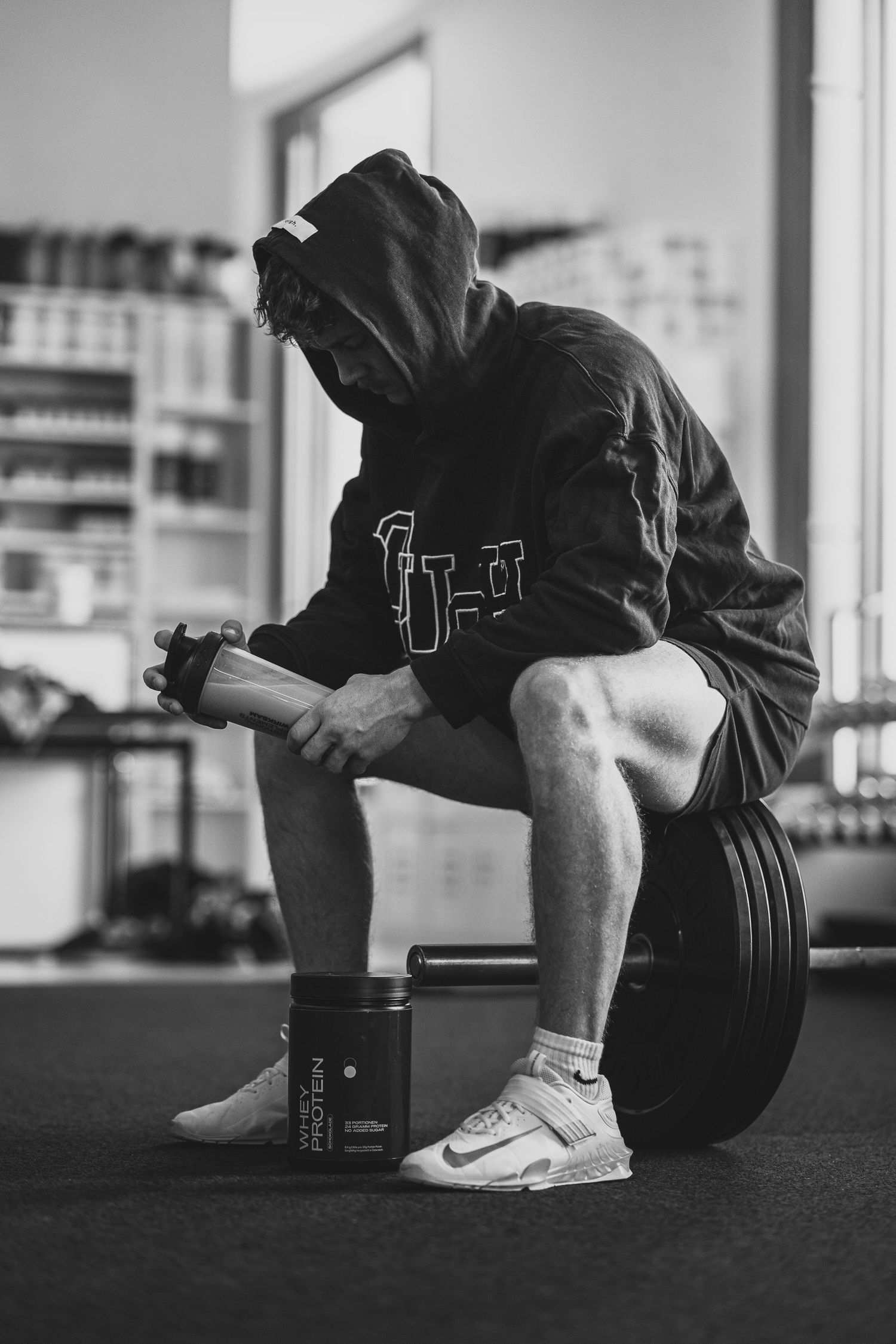 Black and white photo of hooded athlete resting on barbell plates with supplement container nearby.
