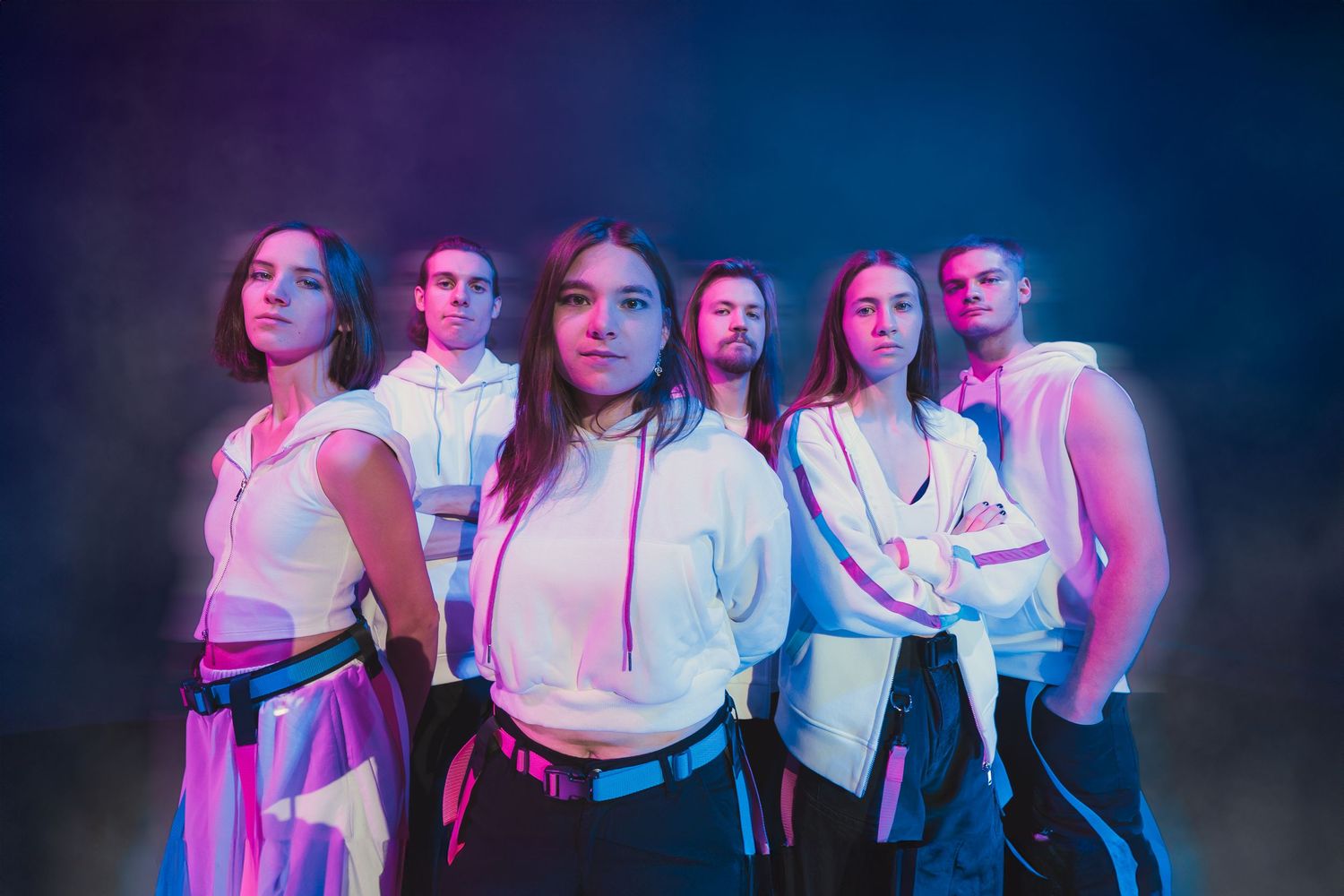 Group of six young dancers posing together under vibrant blue and pink neon studio lighting.