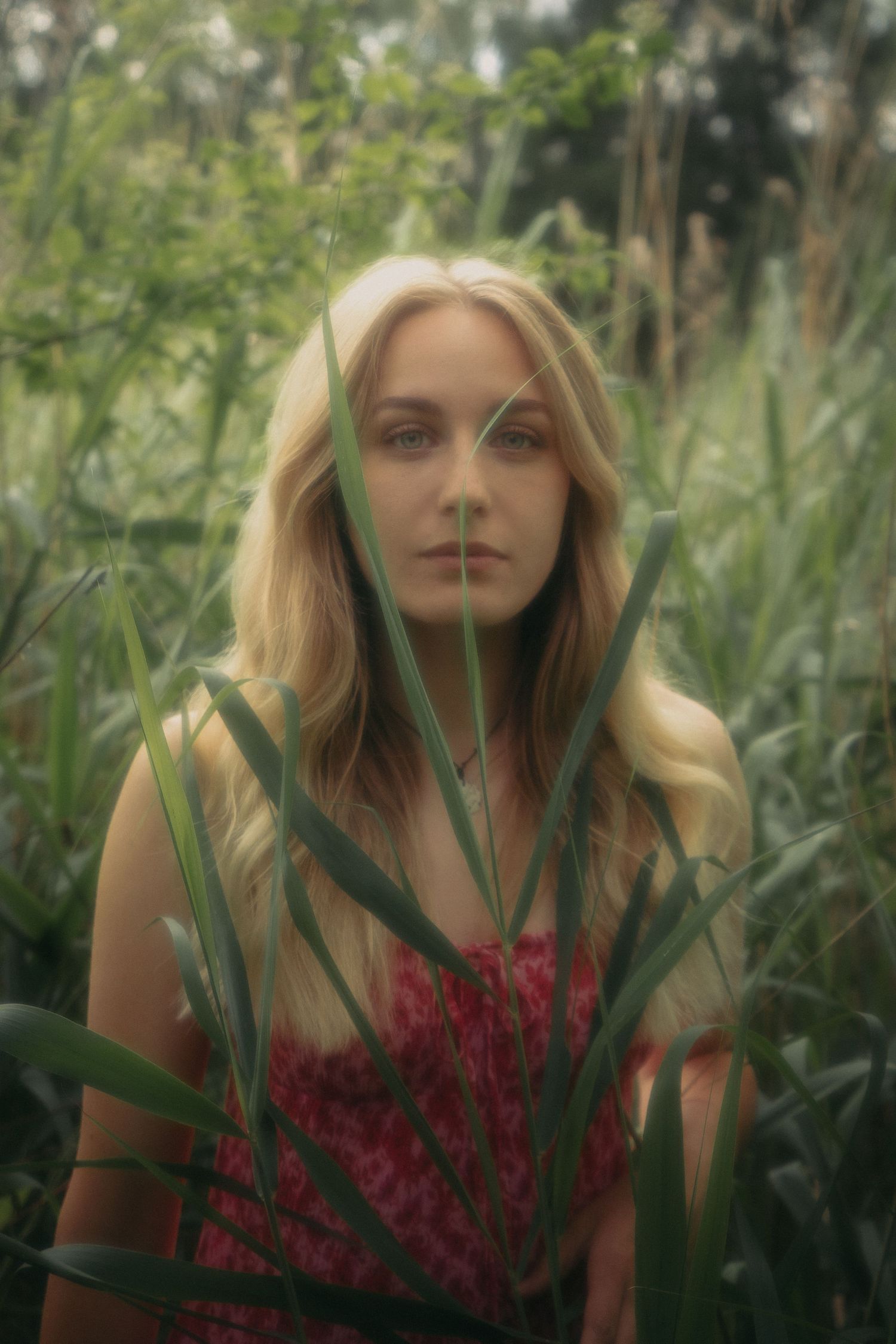 Blonde woman in pink floral top partially hidden behind tall green grass in nature.