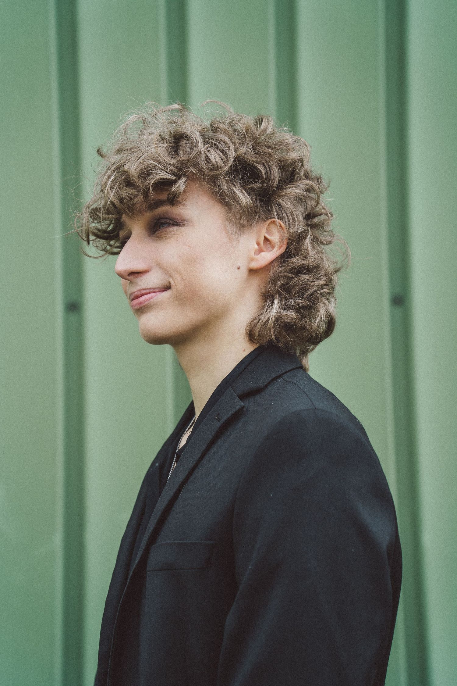 Young person with curly blonde hair wearing black blazer smiling against green wall.