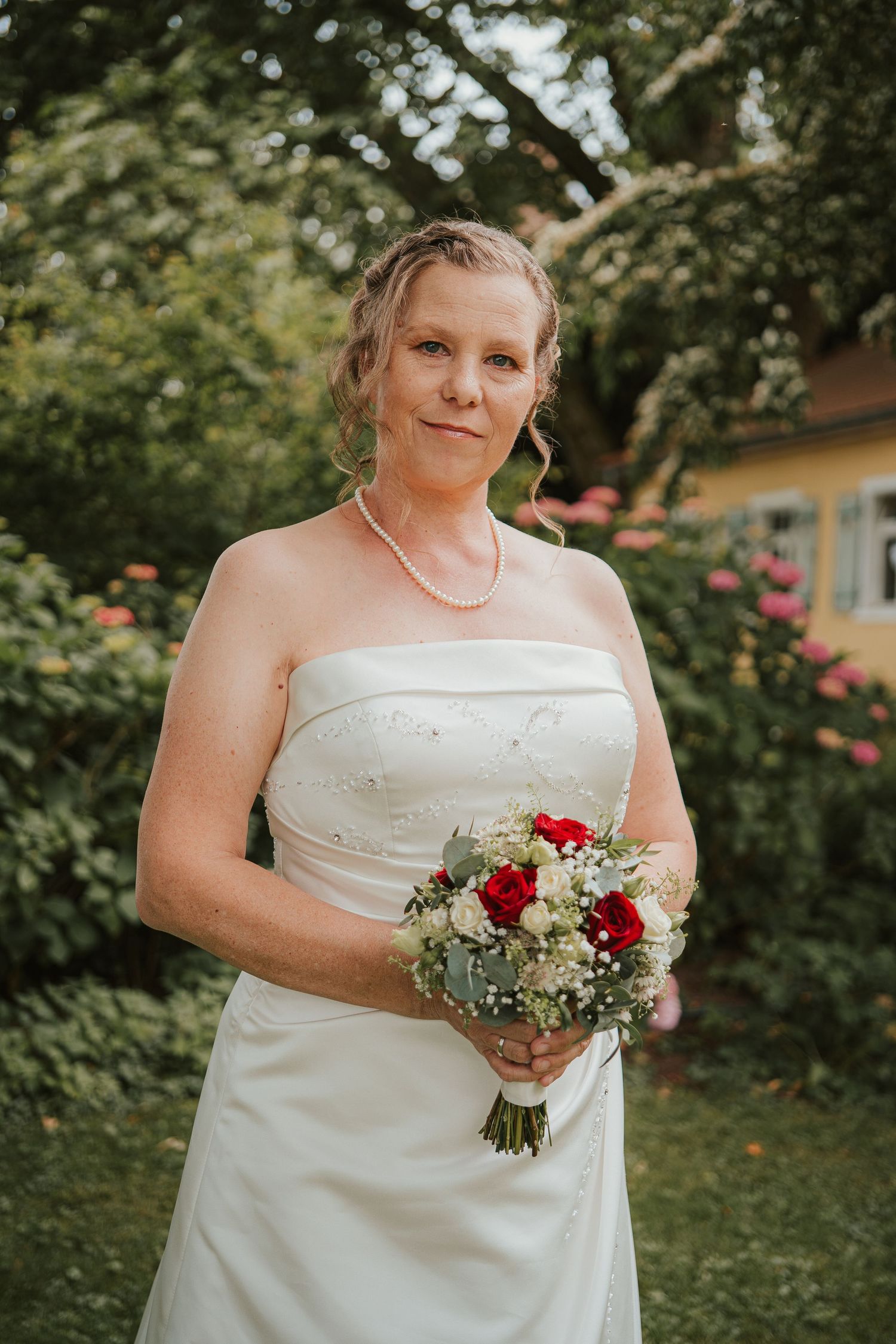 Bride in strapless white wedding gown holding red and white bouquet, posing in a lush garden setting.