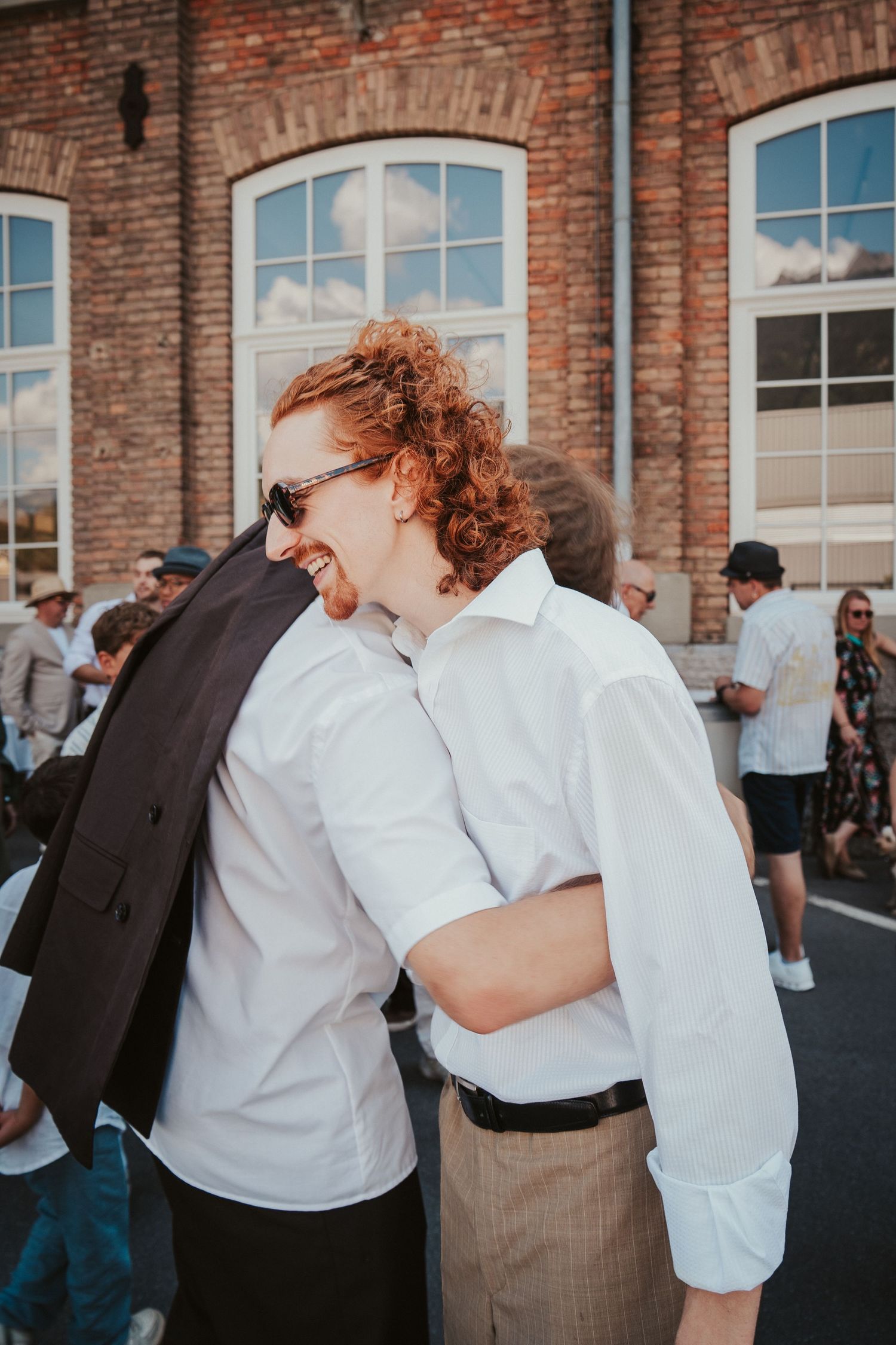 Two red-haired wedding guests share a warm embrace while laughing outside a brick building at a wedding reception.