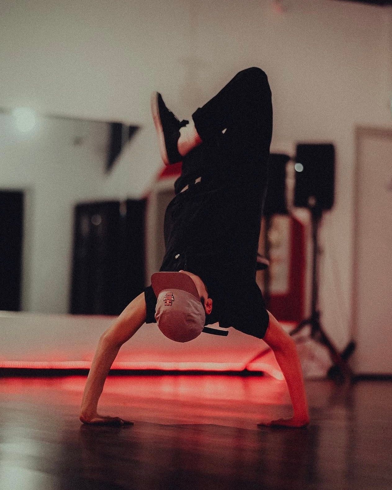 A breakdancer performs a headstand in a dance studio with dramatic red lighting reflecting off the floor.