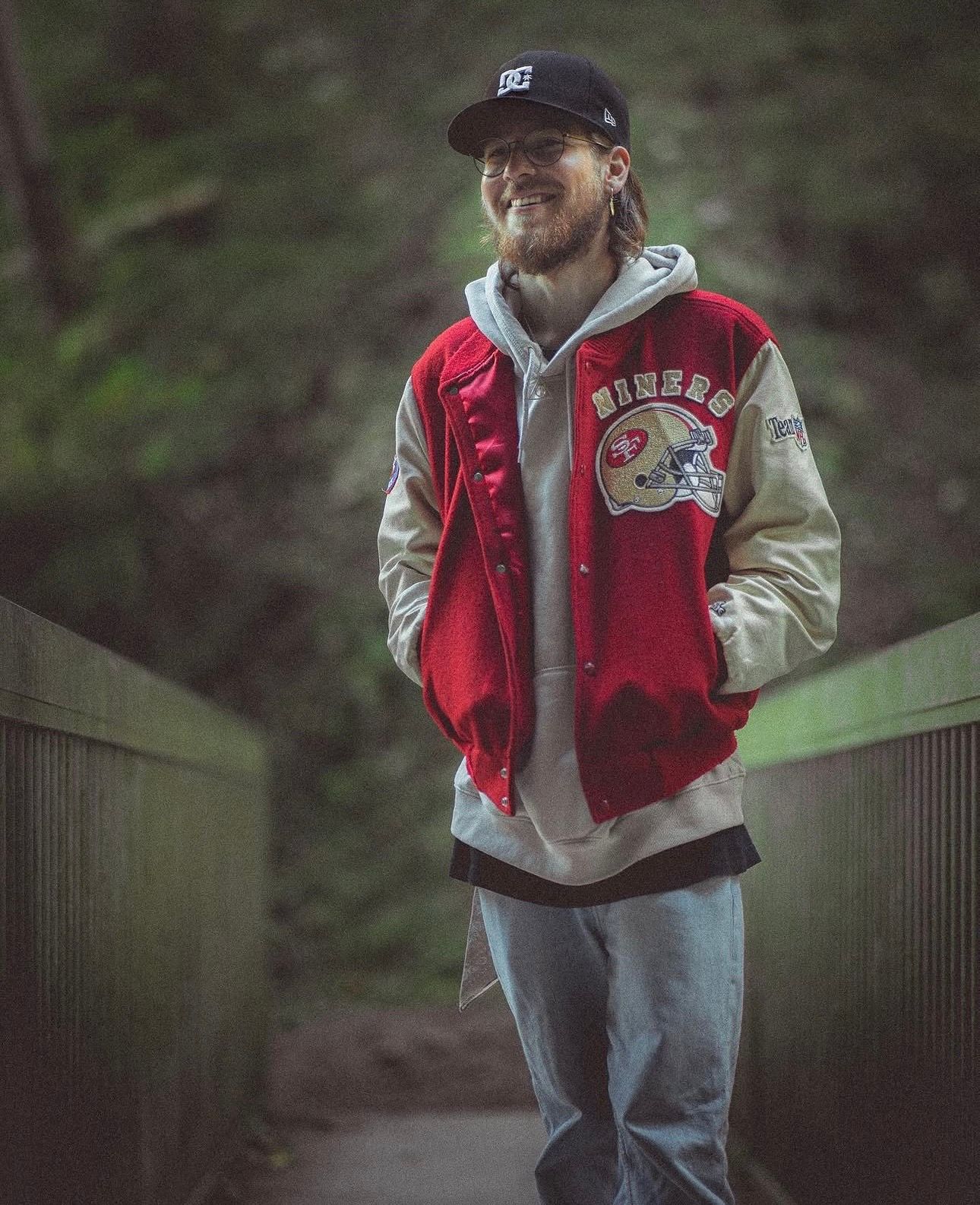 Smiling man wearing San Francisco 49ers varsity jacket and cap standing on forest bridge path.
