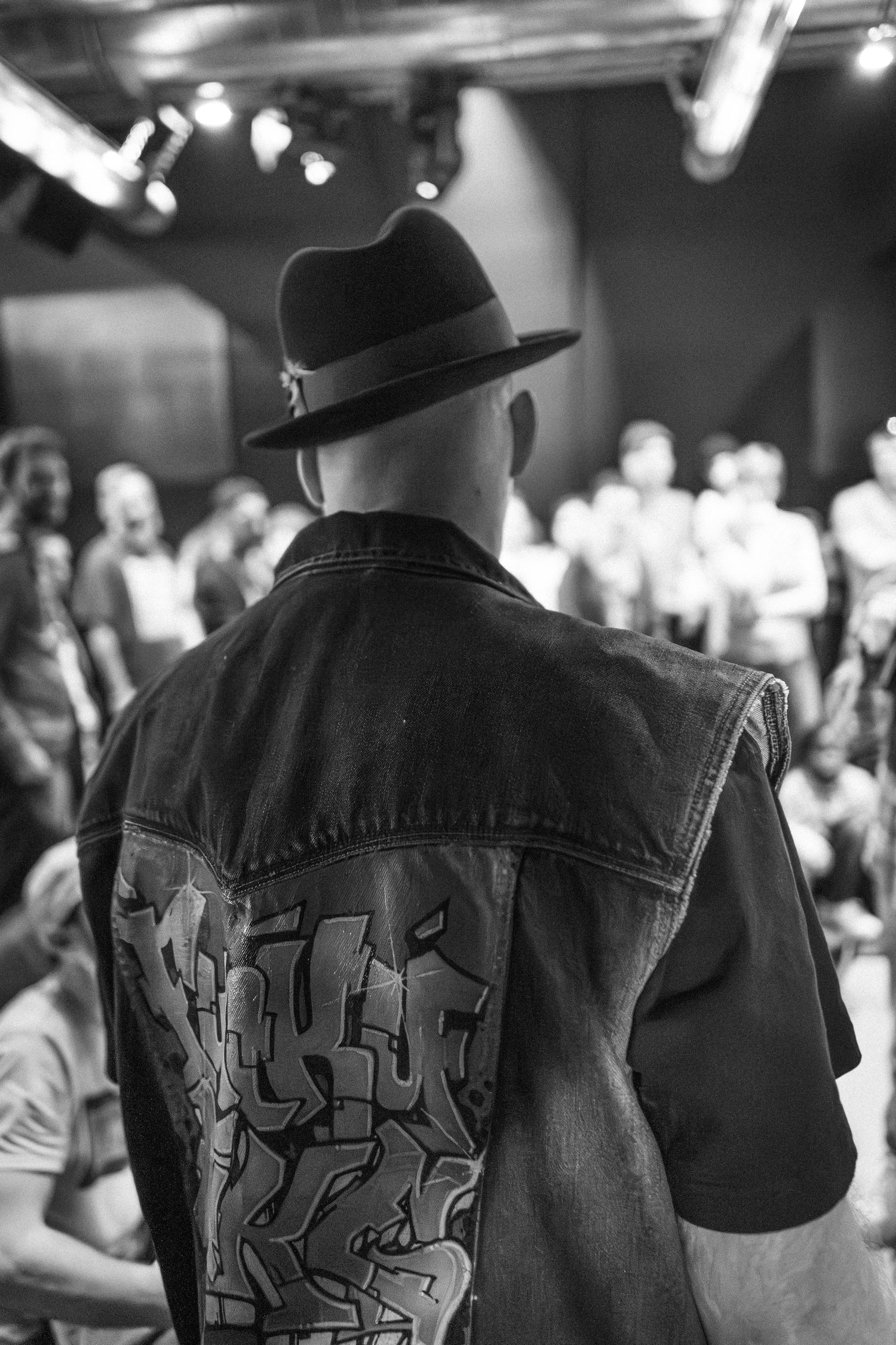 Man in a fedora and graffiti denim vest stands before a crowd at a hip-hop event.