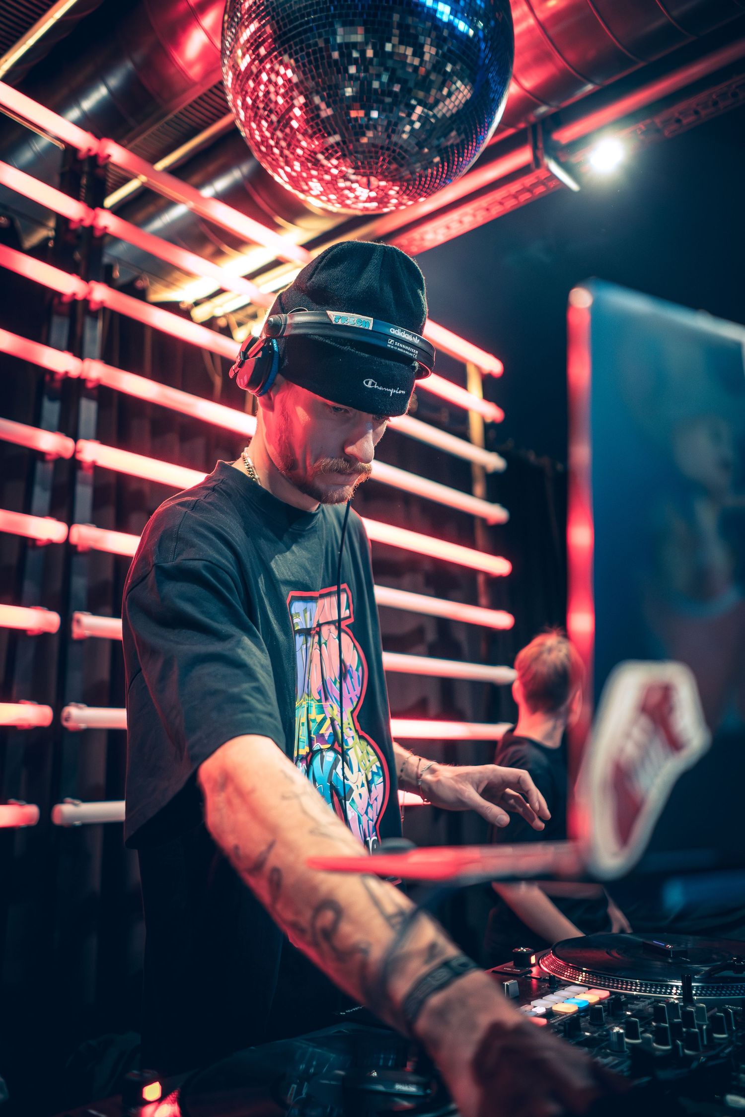 Tattooed DJ with a Champion cap mixes tracks under a disco ball at a neon-lit hip-hop event.