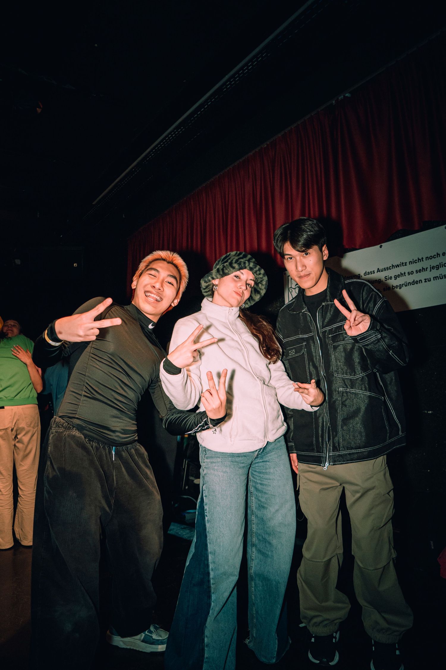 Three young people pose together making peace signs in a dimly lit venue with a red curtain backdrop.