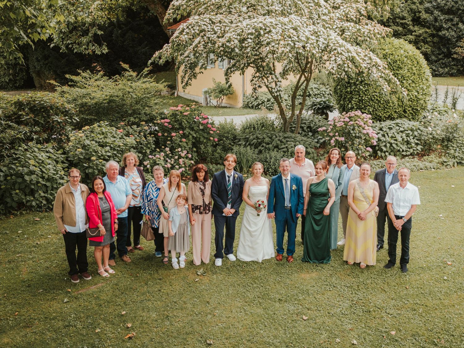 Wedding party group photo on green lawn with blooming white tree and yellow house backdrop.