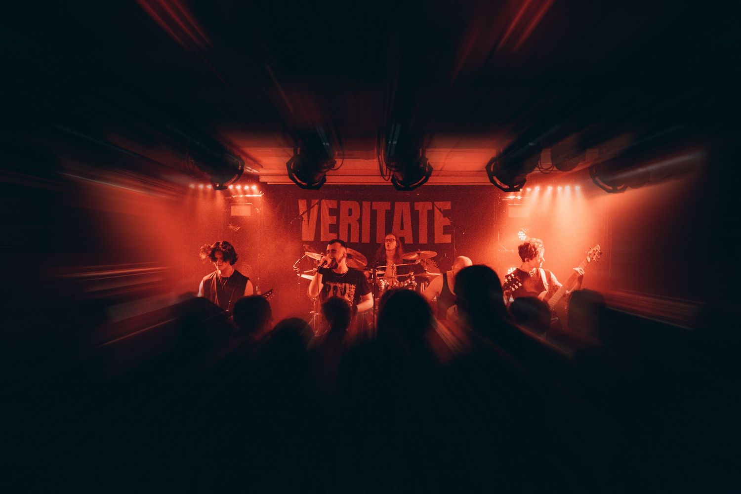 Band performing on stage under red lights with VERITATE sign backdrop.