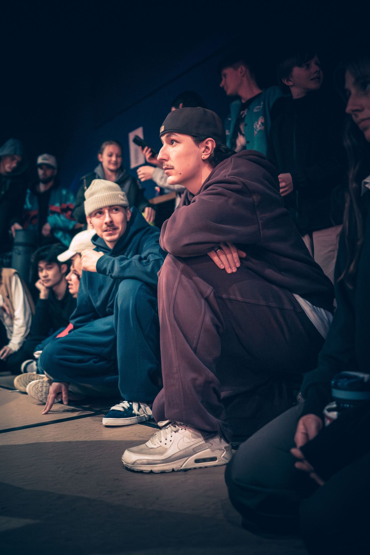 Two breakdancers in streetwear crouch and watch a battle at an urban hip-hop dance event.