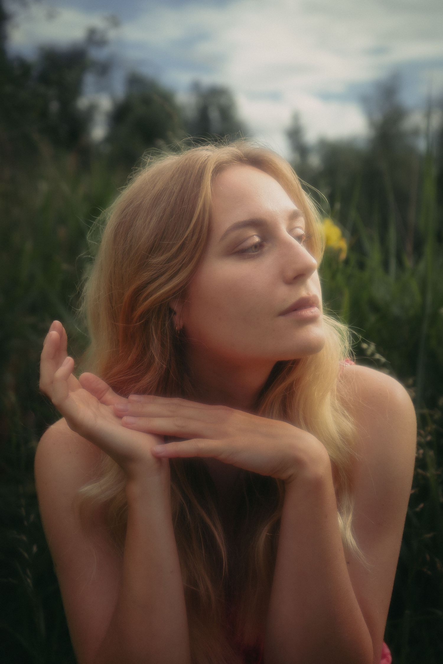 Blonde woman with eyes closed holding hands under chin sitting in lush green field.