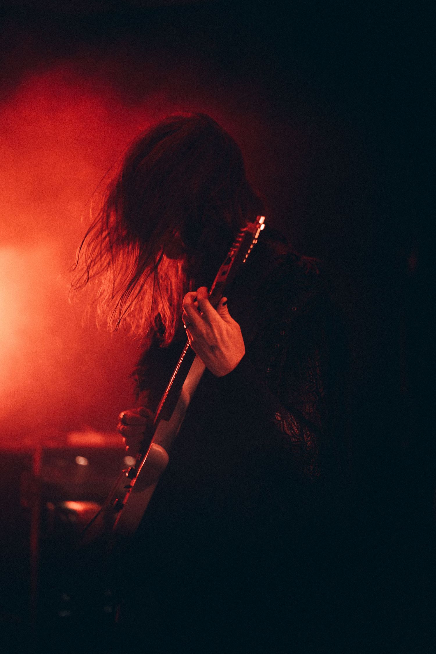 Long-haired guitarist playing electric guitar in moody red smoke-filled stage light.