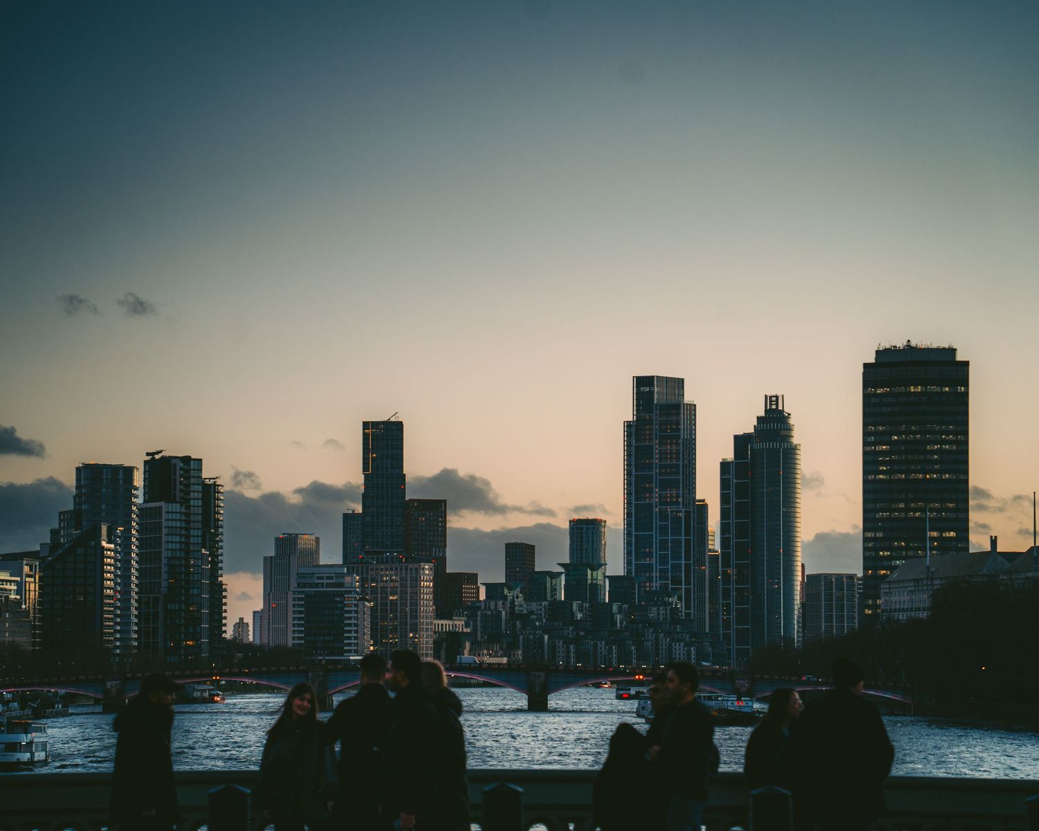 Silhouette of people watching city skyline at dusk with skyscrapers reflected over river.