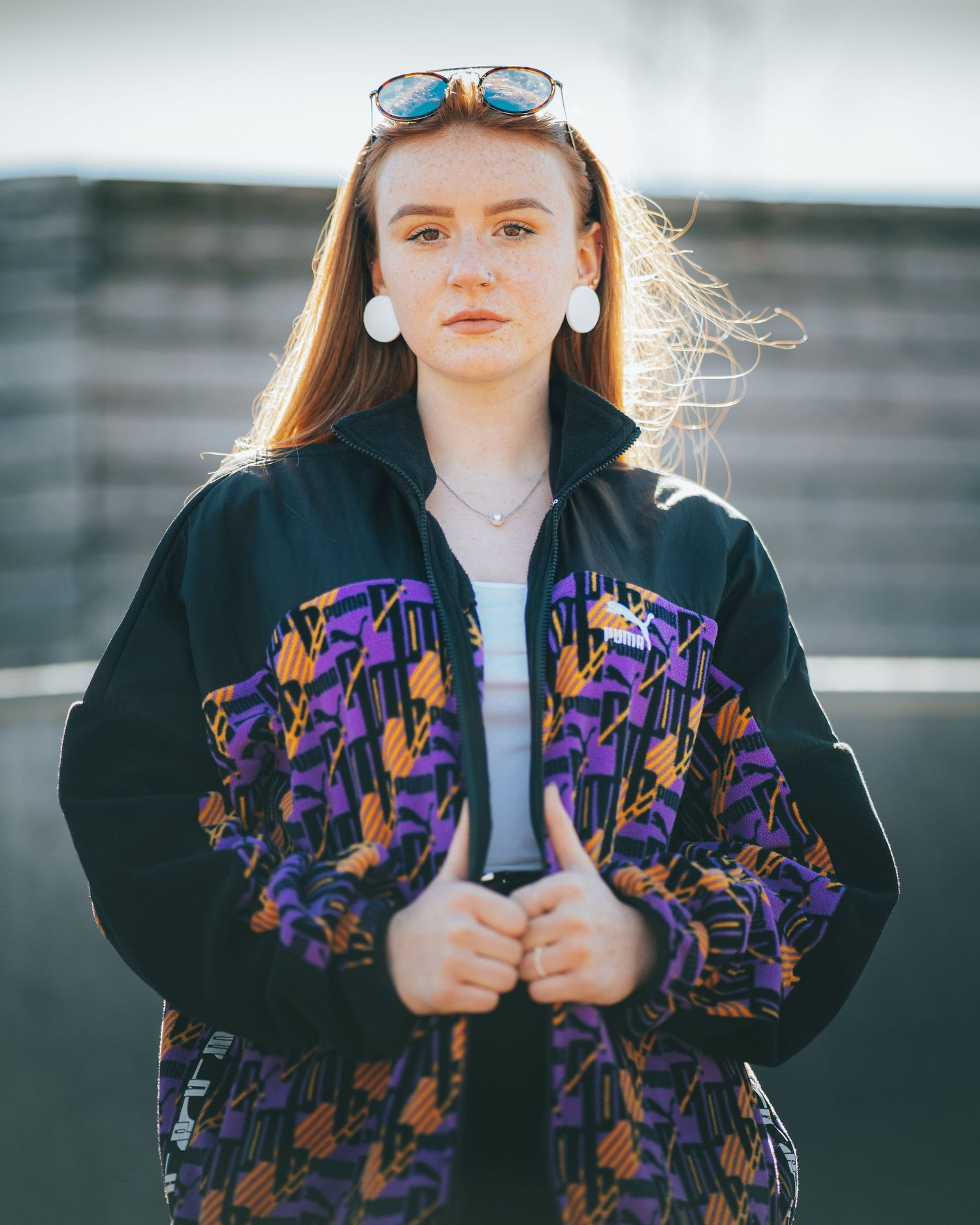 Young redhead woman in colorful vintage jacket with white earrings and sunglasses outdoors.