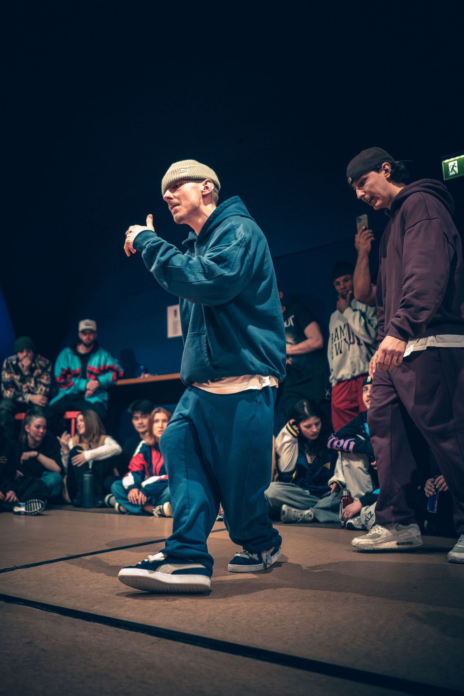 A breakdancer in a blue jacket and beanie performs footwork moves on a dance floor before a live audience.