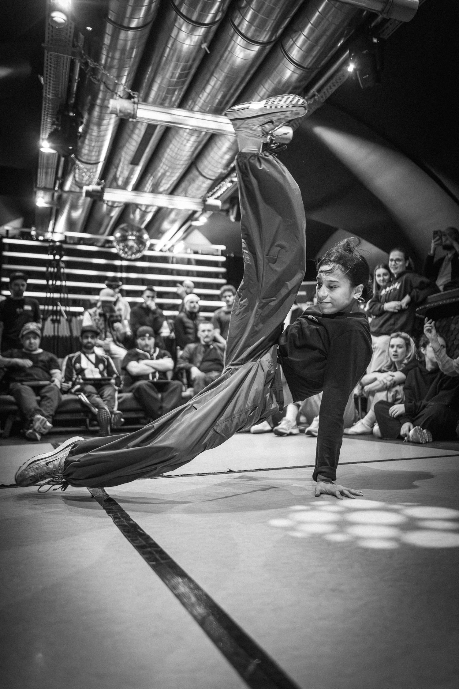 Black and white photo of a young breakdancer performing a dynamic freeze move at a dance battle.