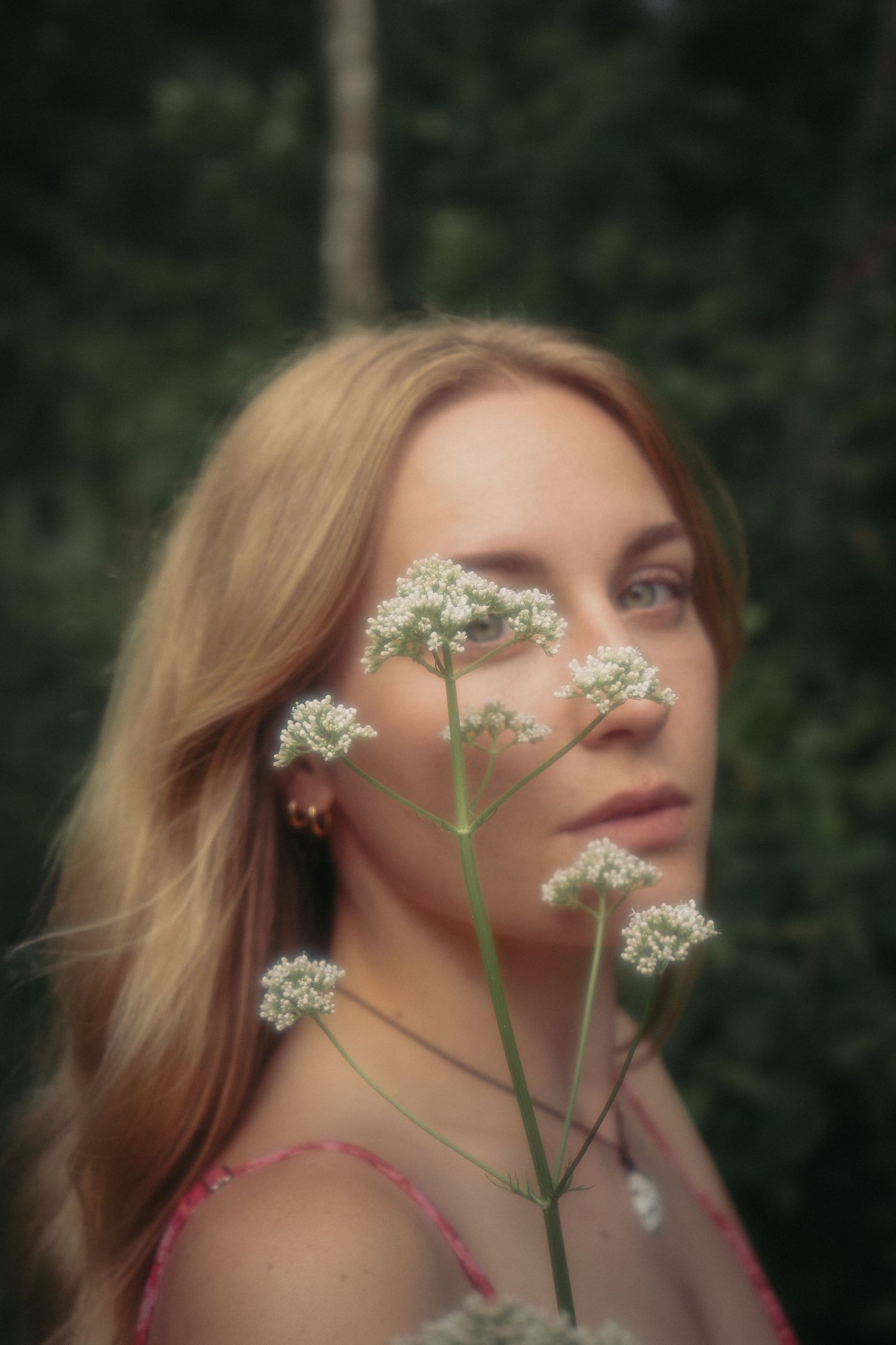 Young blonde woman gazing upward with delicate white wildflowers in the foreground against a blurred forest backdrop.