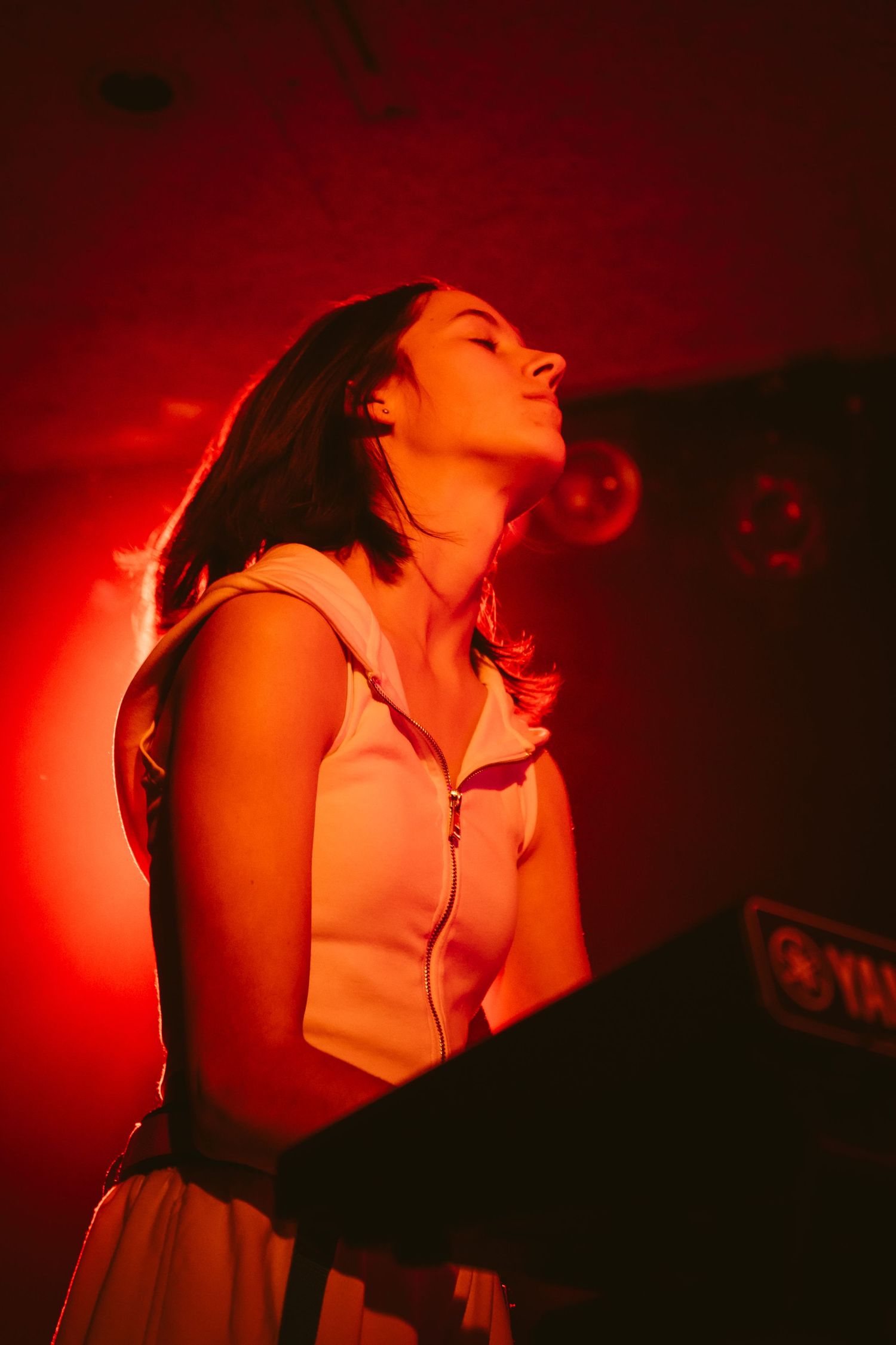 Female keyboardist performing on stage bathed in dramatic red concert lighting with eyes closed.