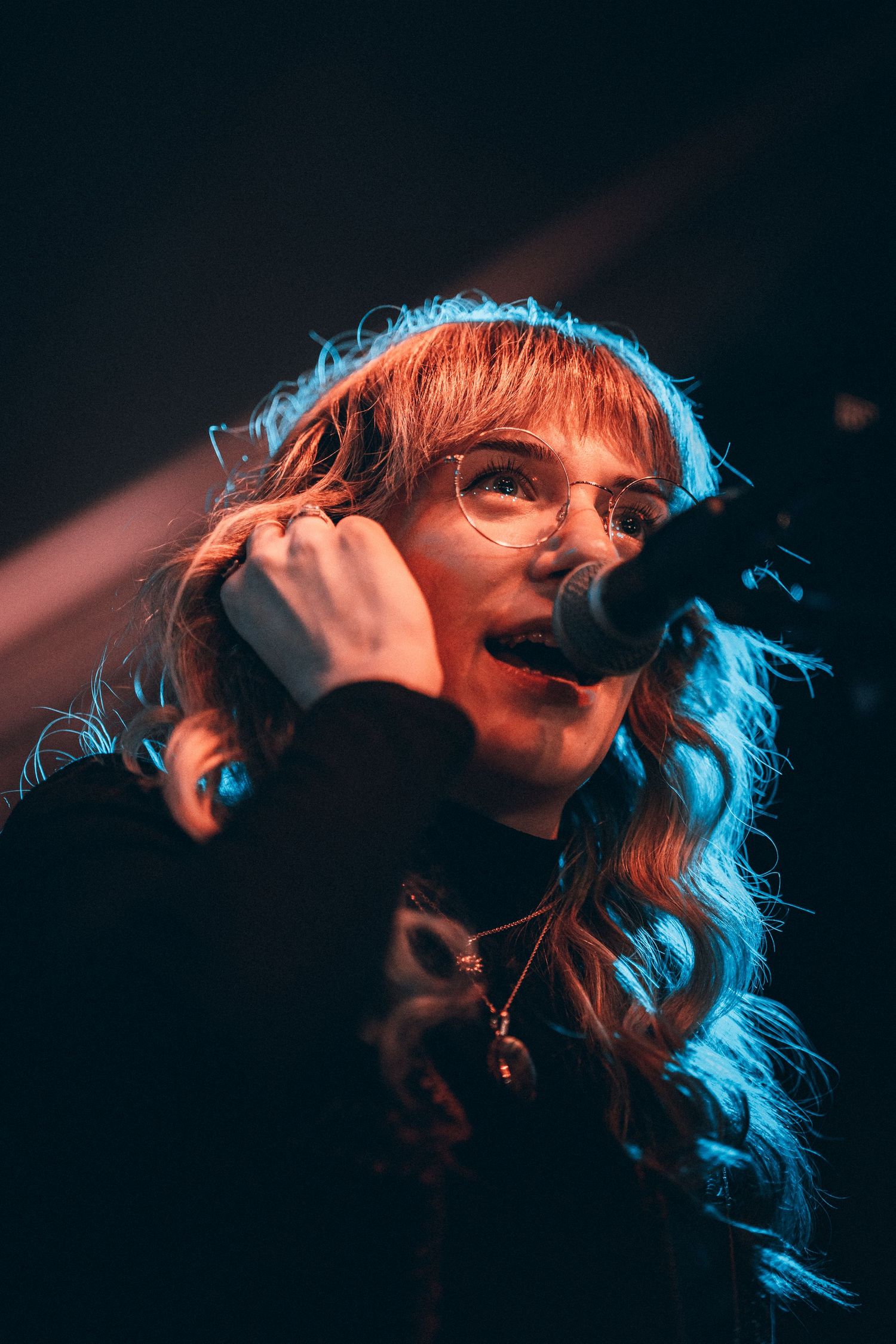 Female vocalist with curly red hair and glasses singing into microphone on stage.