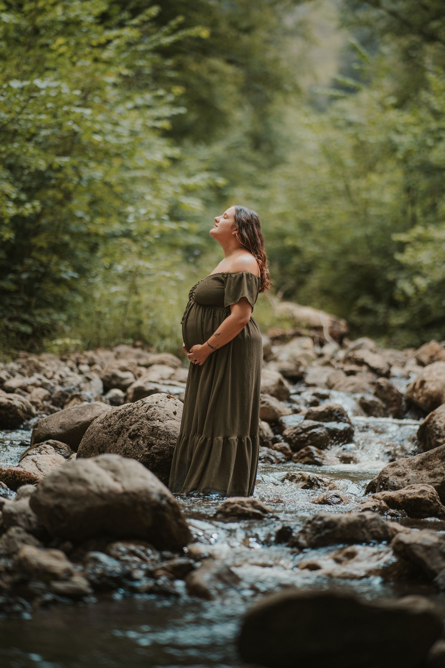 Pregnant woman in olive green dress standing in a rocky stream surrounded by lush forest.