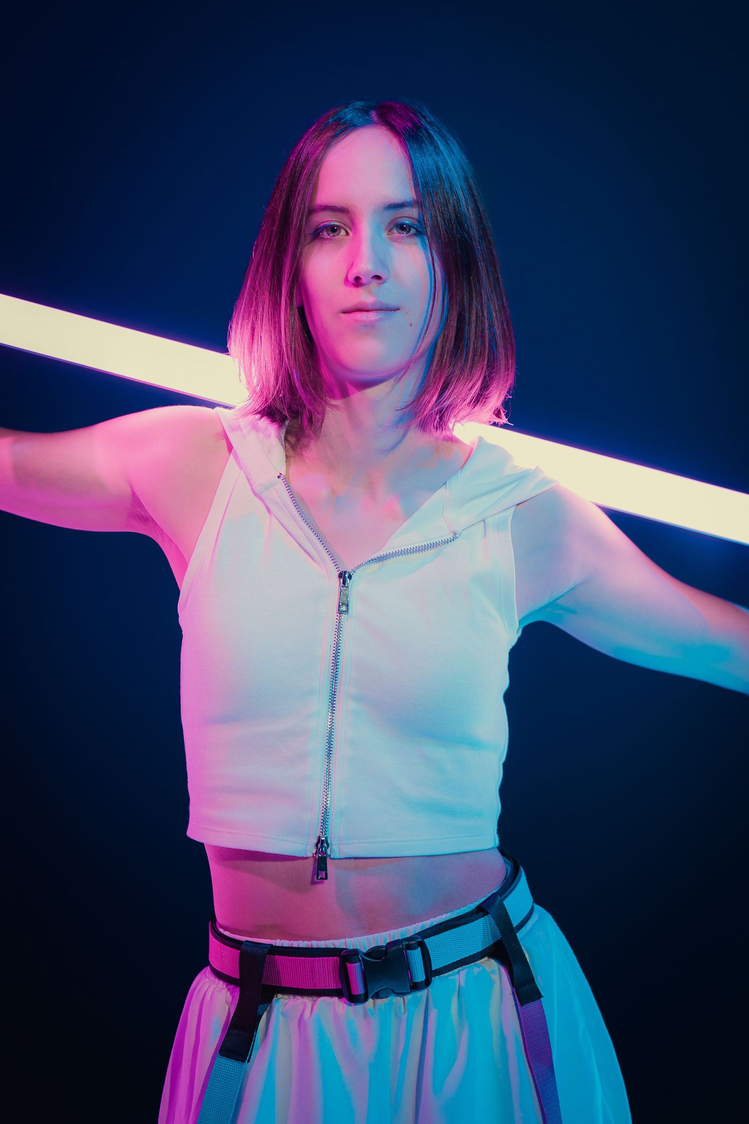 Young woman in white crop top posing with neon light tube in blue and pink studio.