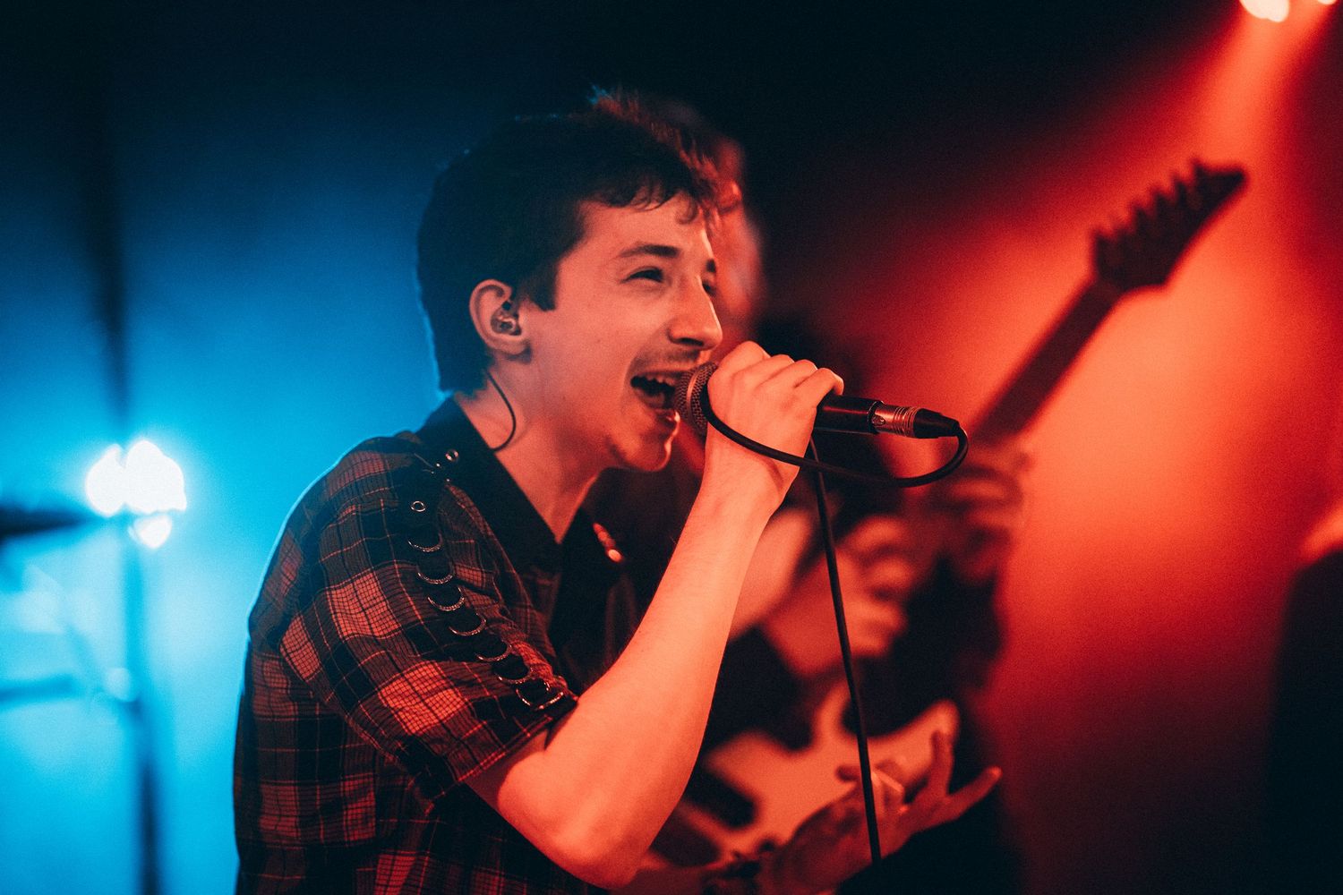 Young vocalist singing into microphone under blue and red concert stage lighting.