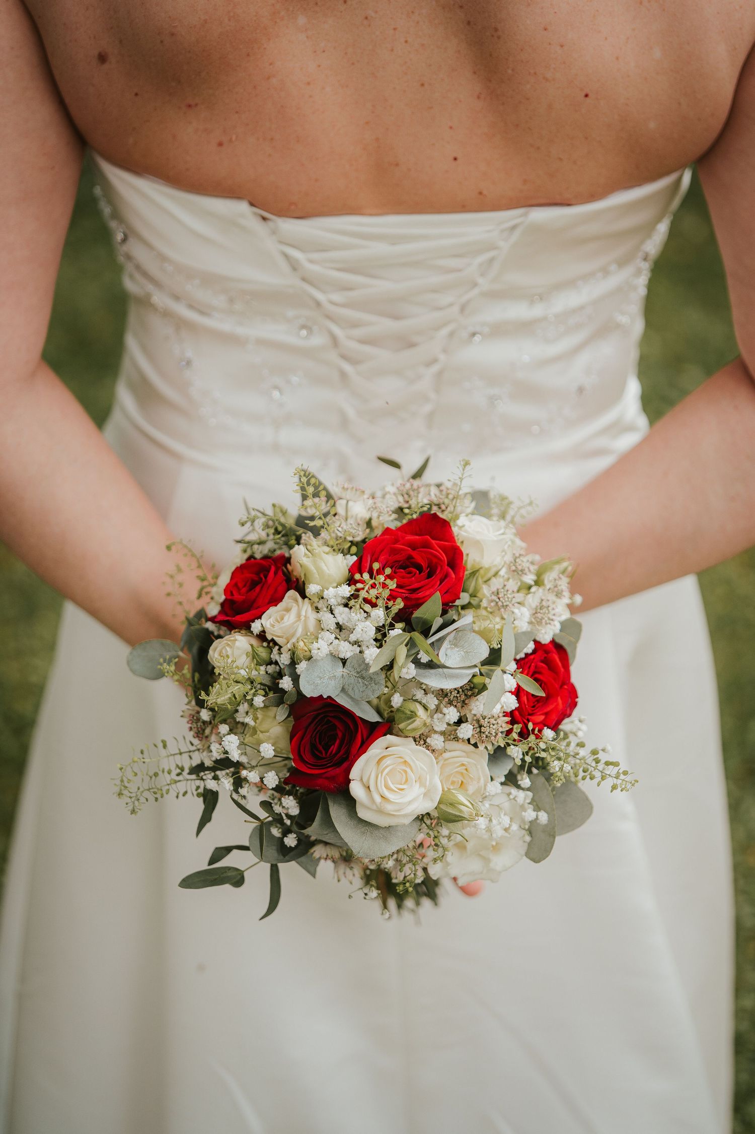 Bride in a lace-back white wedding gown holding a bouquet of red and white roses with eucalyptus greenery.