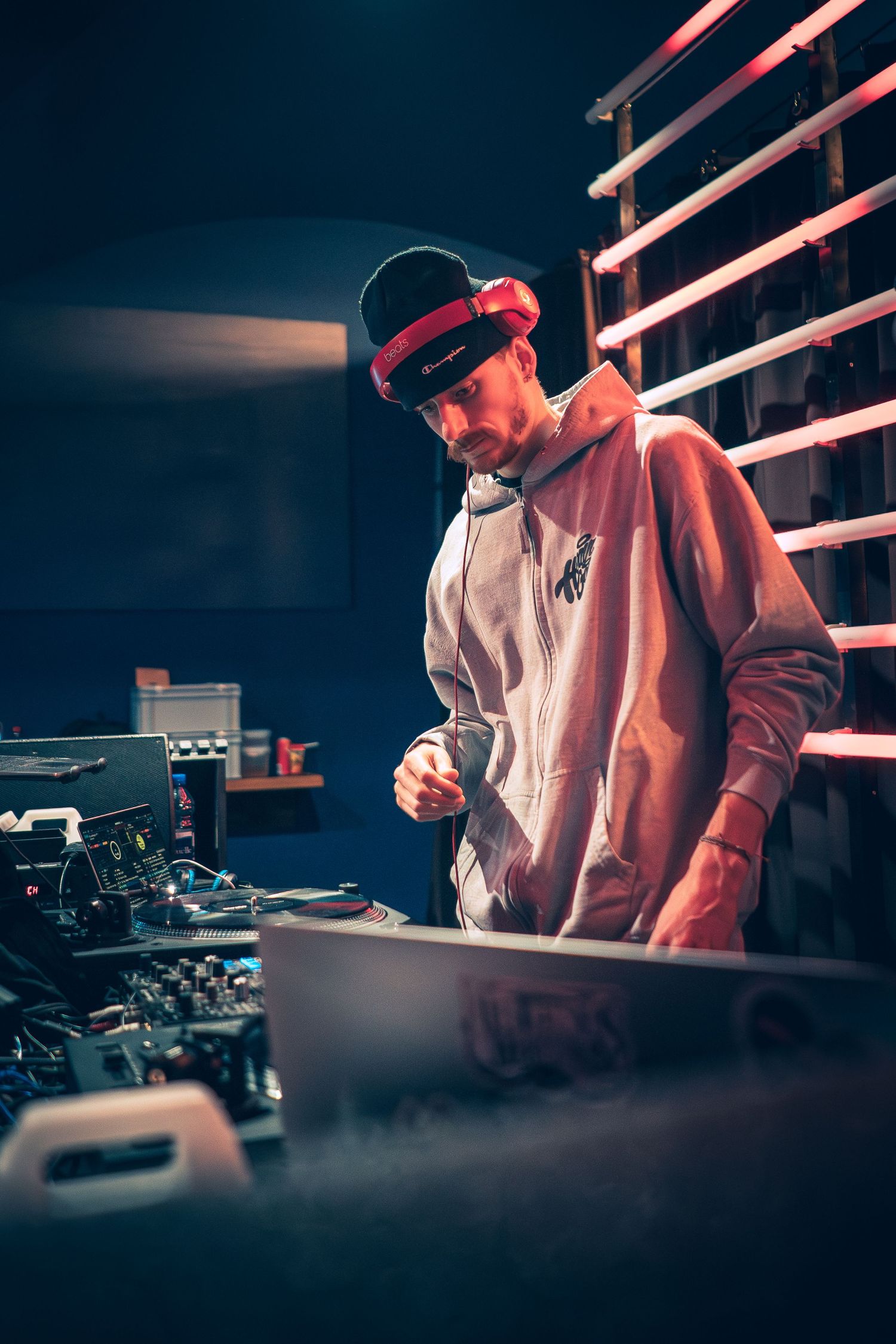 A DJ in a hoodie and cap focuses on mixing equipment in a moody blue and red lit studio space.