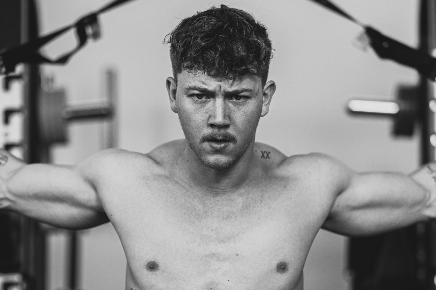 Black and white close-up of shirtless muscular man performing cable exercise with intense expression.