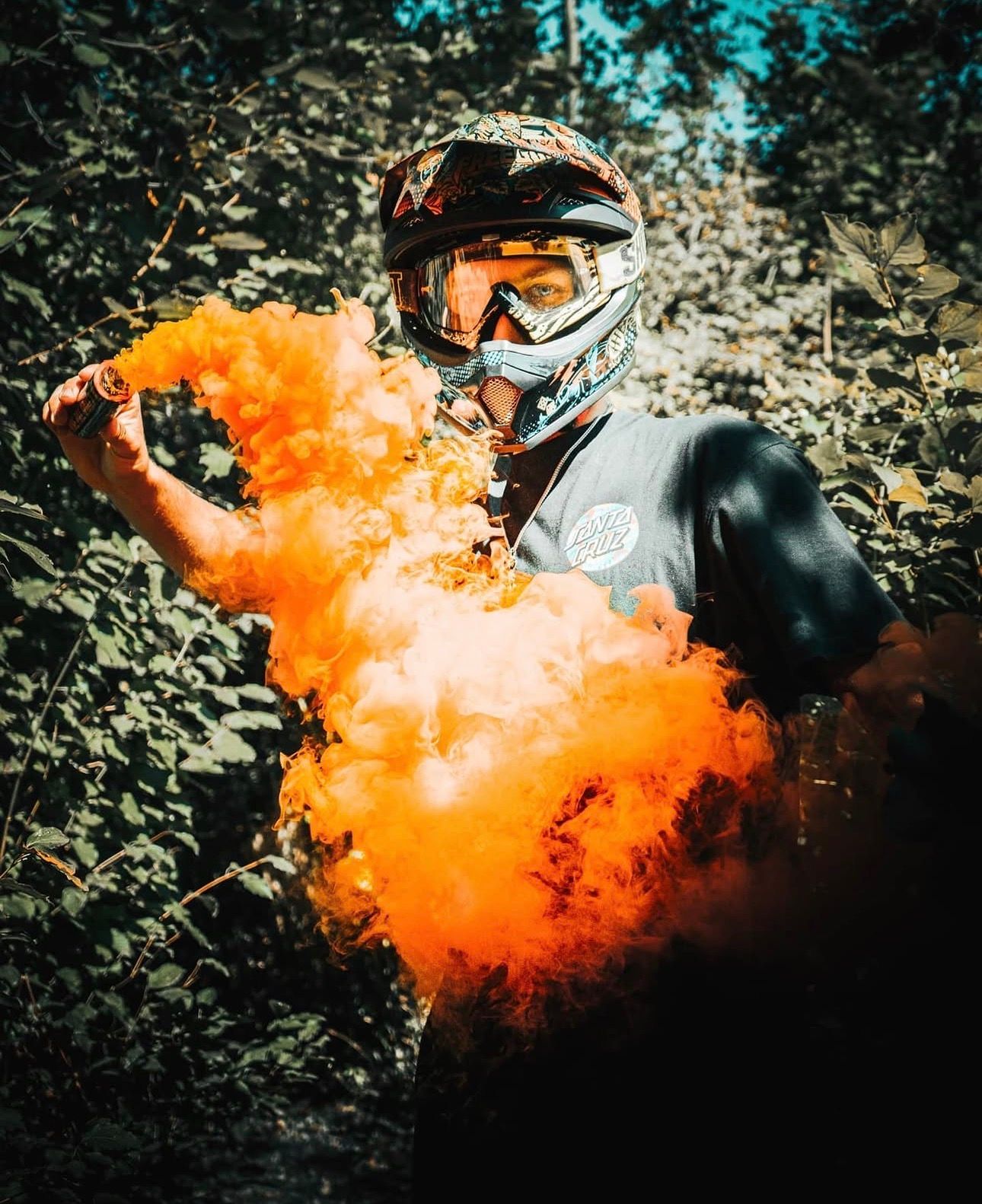 Person in motocross helmet holding vibrant orange smoke bomb surrounded by forest greenery.