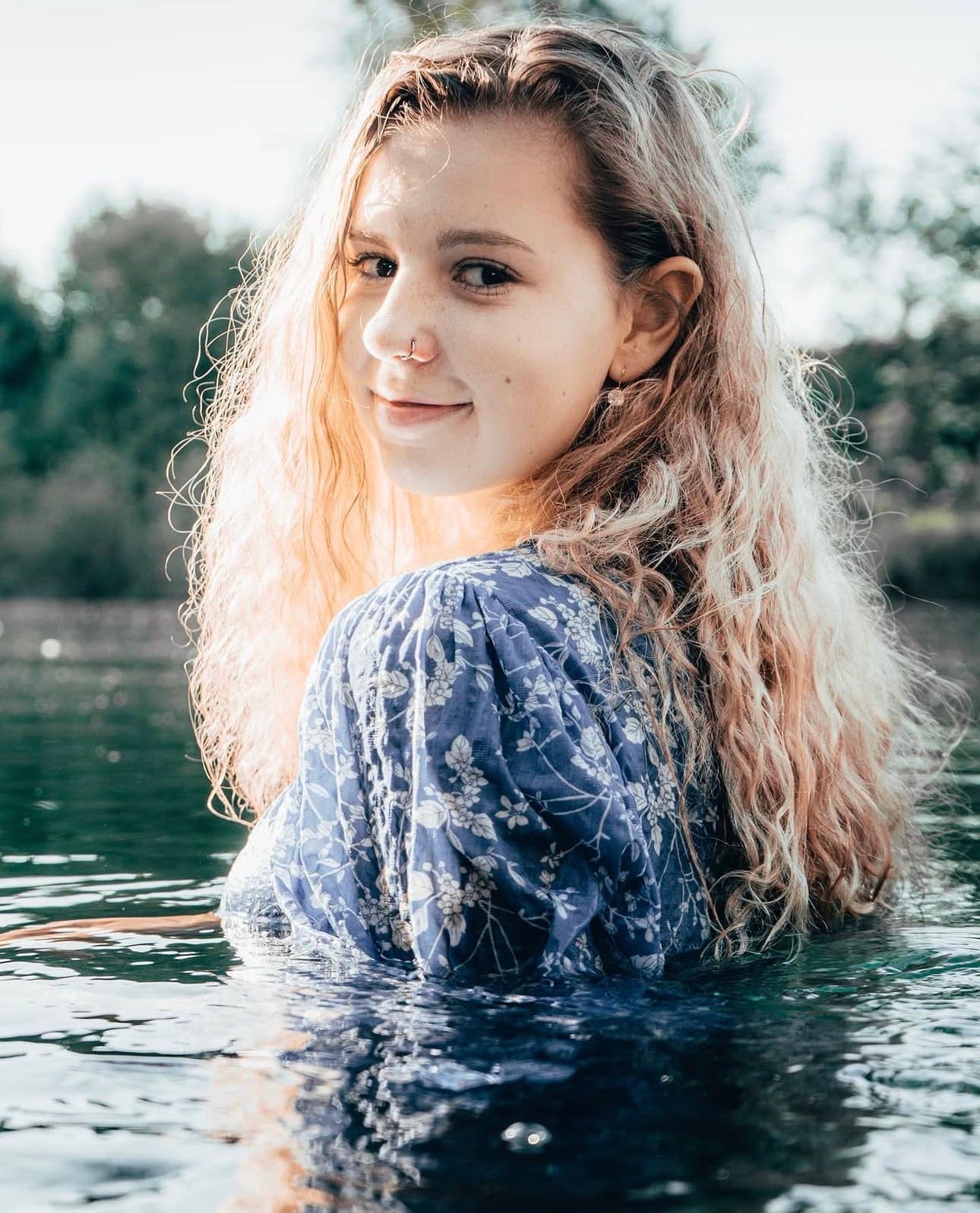 Smiling young woman with blonde hair standing in a lake wearing a blue floral top outdoors.