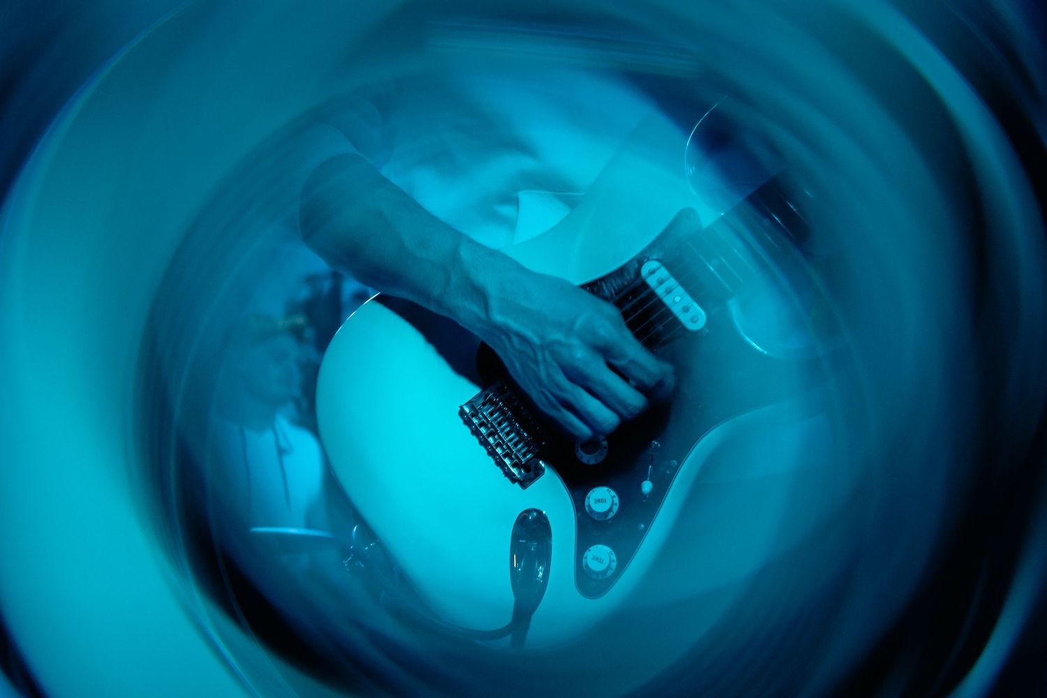 Close-up fisheye view of guitarist playing electric guitar under blue stage lighting.