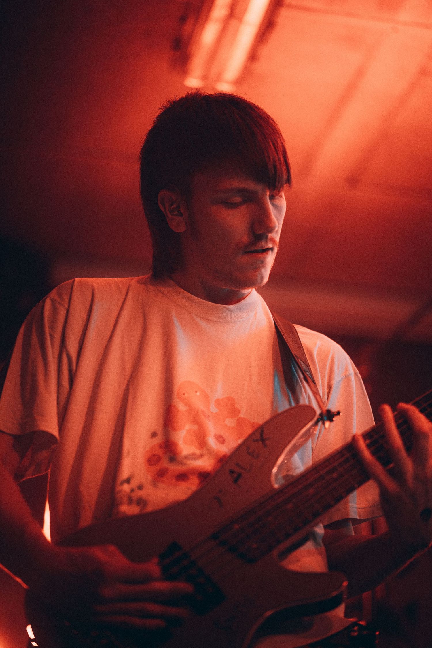 Guitarist in white t-shirt performing under red stage lighting at a live music venue.