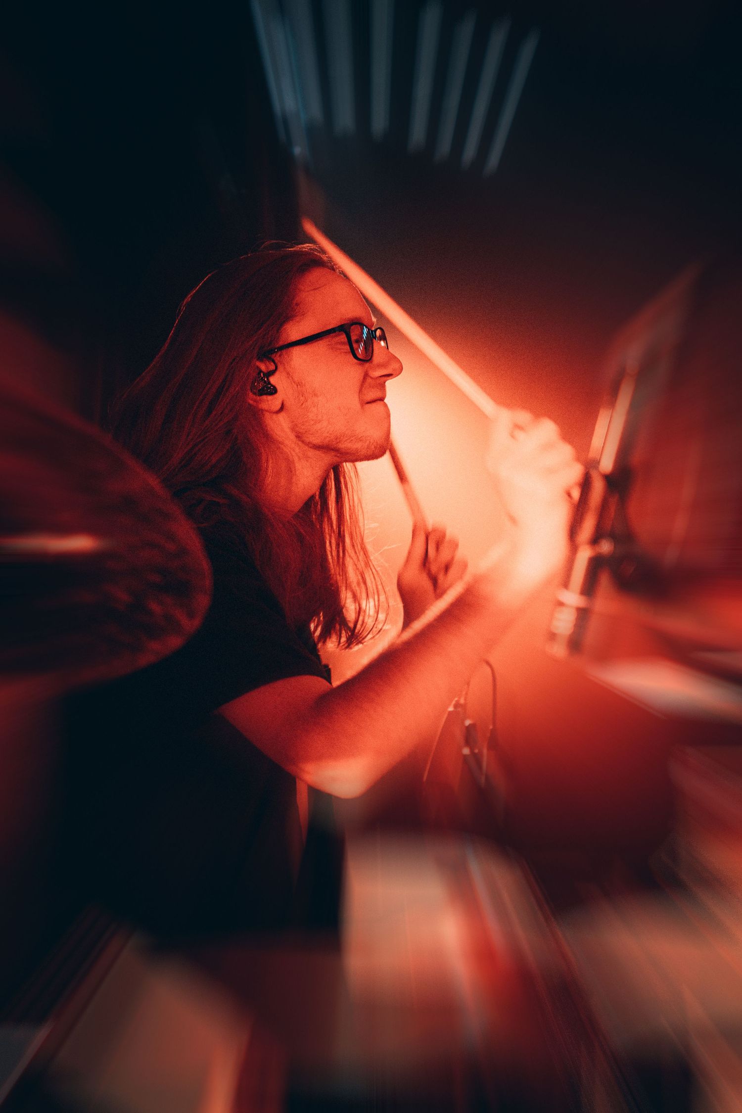 Female drummer with glasses playing drums under dramatic red stage lighting.