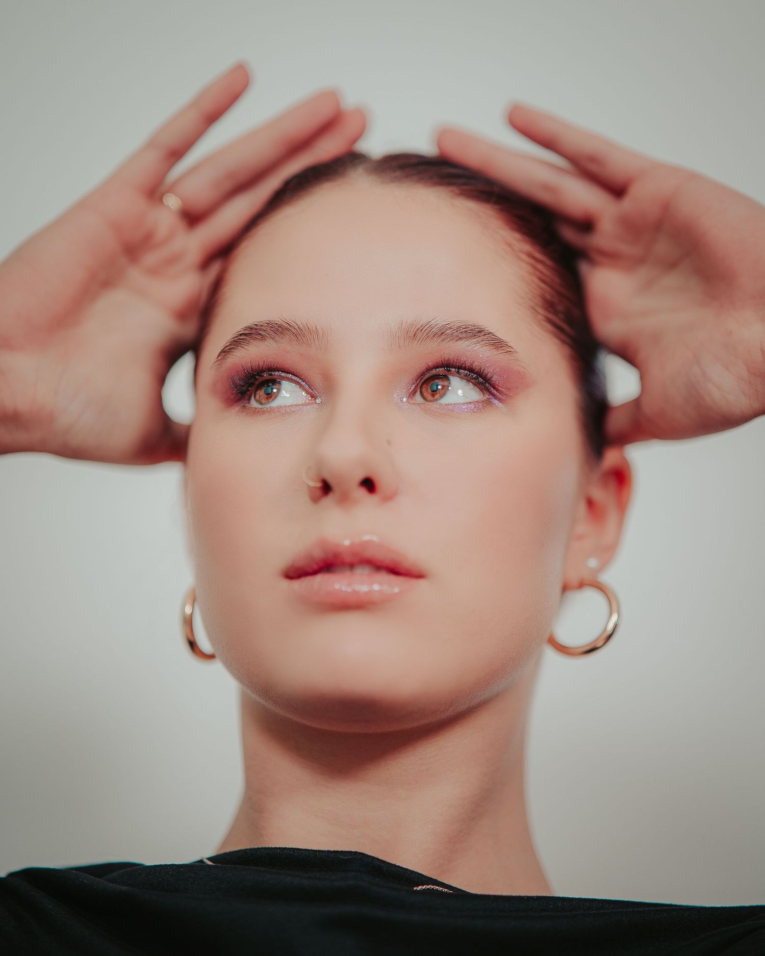 Young woman with hoop earrings holding hands above her head gazing upward against neutral background.