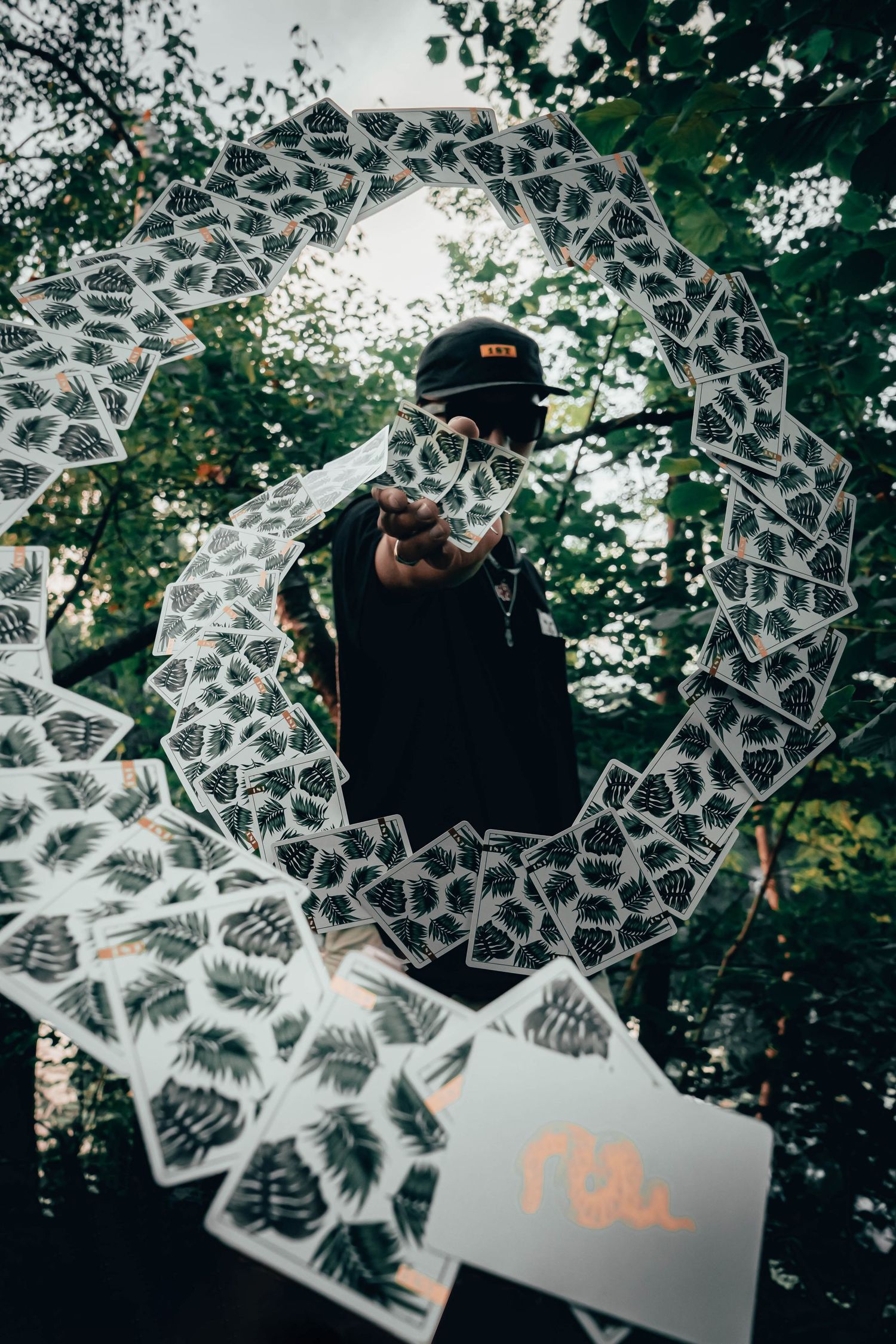 Man pointing at camera surrounded by a circular arrangement of playing cards in a forest setting.