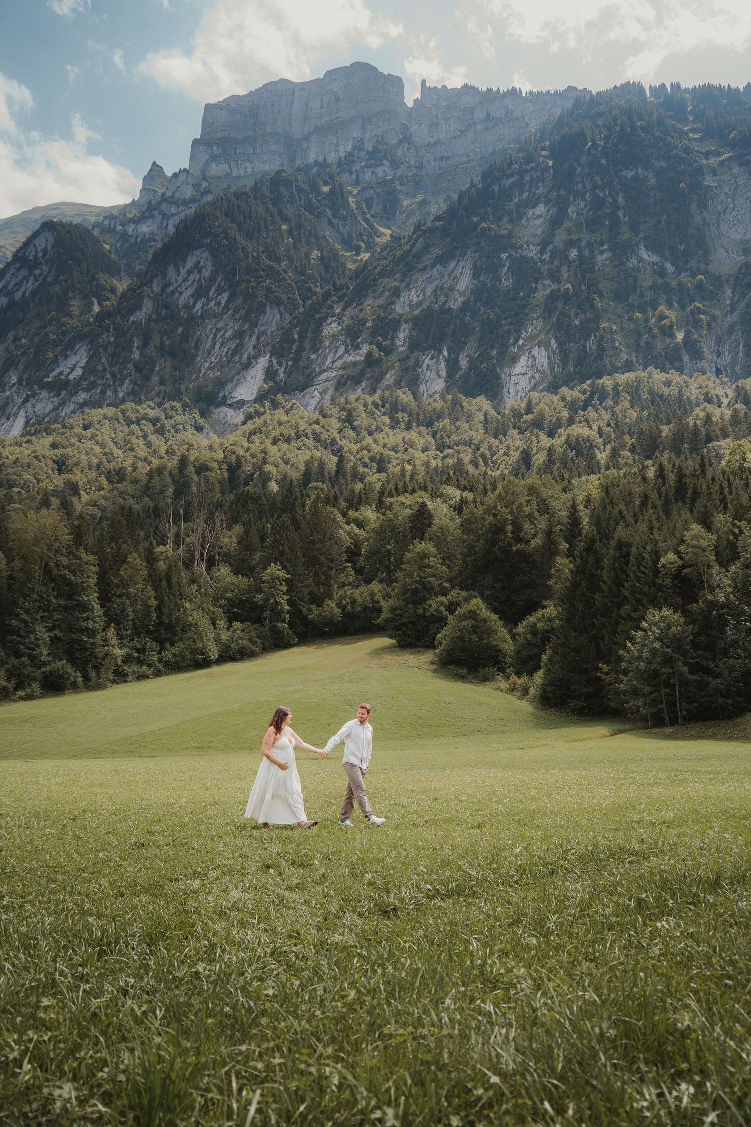 Couple in wedding attire holding hands in a lush green alpine meadow with dramatic mountain cliffs in the background.