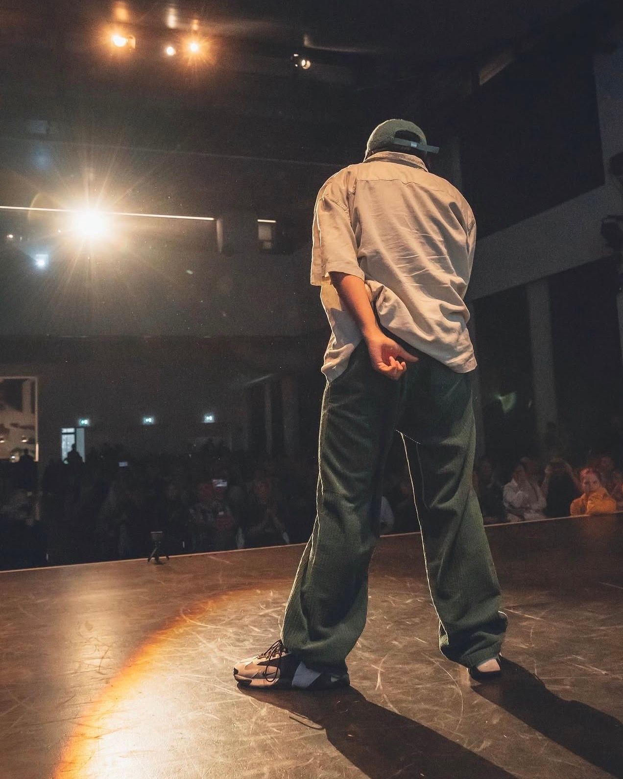 A performer stands with their back to the audience on a lit stage wearing a cap and wide-leg green trousers.